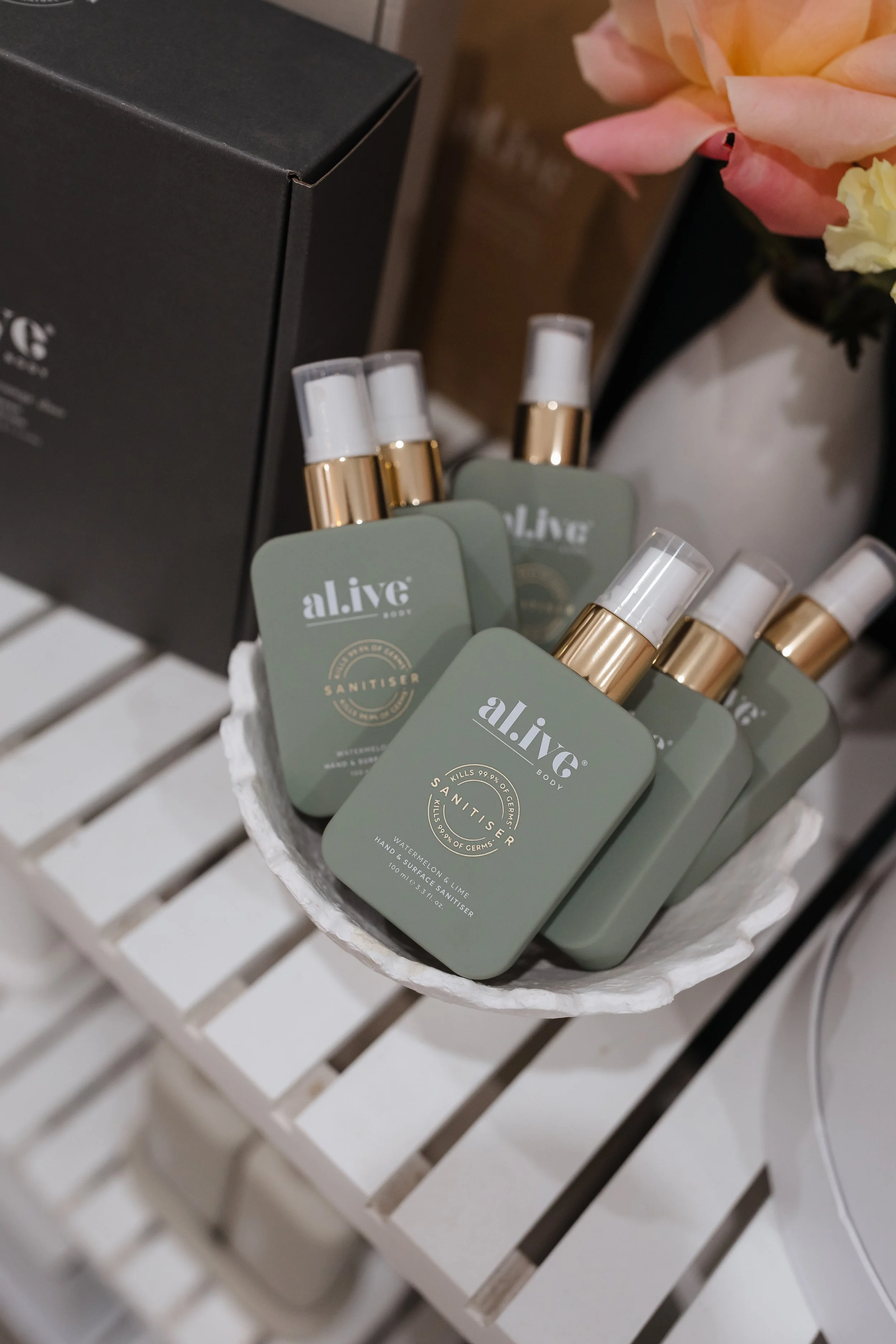 A basket of green soap bottles labeled 'alive' placed on a white slatted surface, with a black box and pink and yellow flowers in the background.