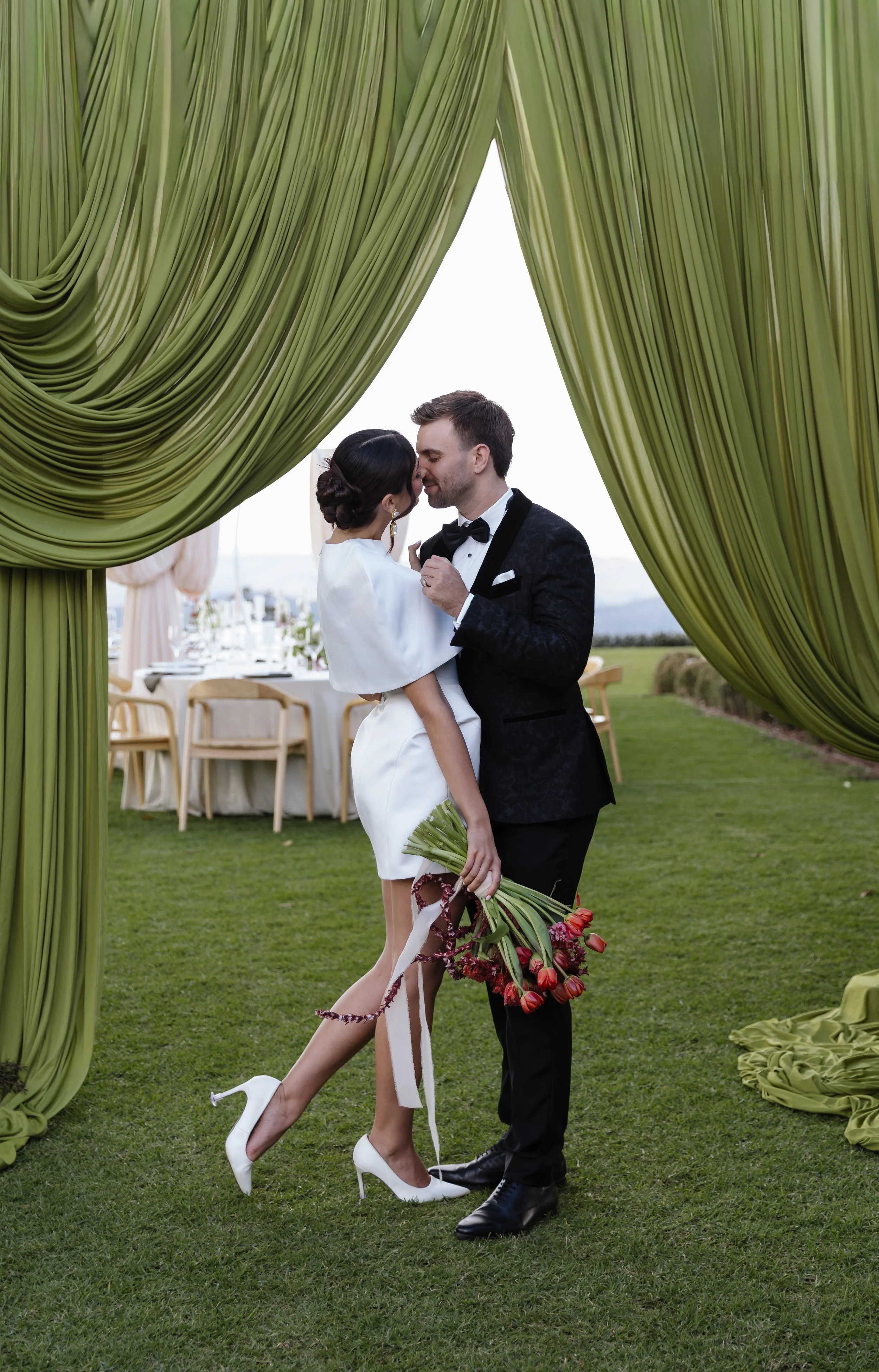 A bride and groom share a kiss under green curtains at an outdoor wedding reception. The bride is holding a bouquet of red and pink tulips, and the groom is wearing a black tuxedo with a bow tie.
