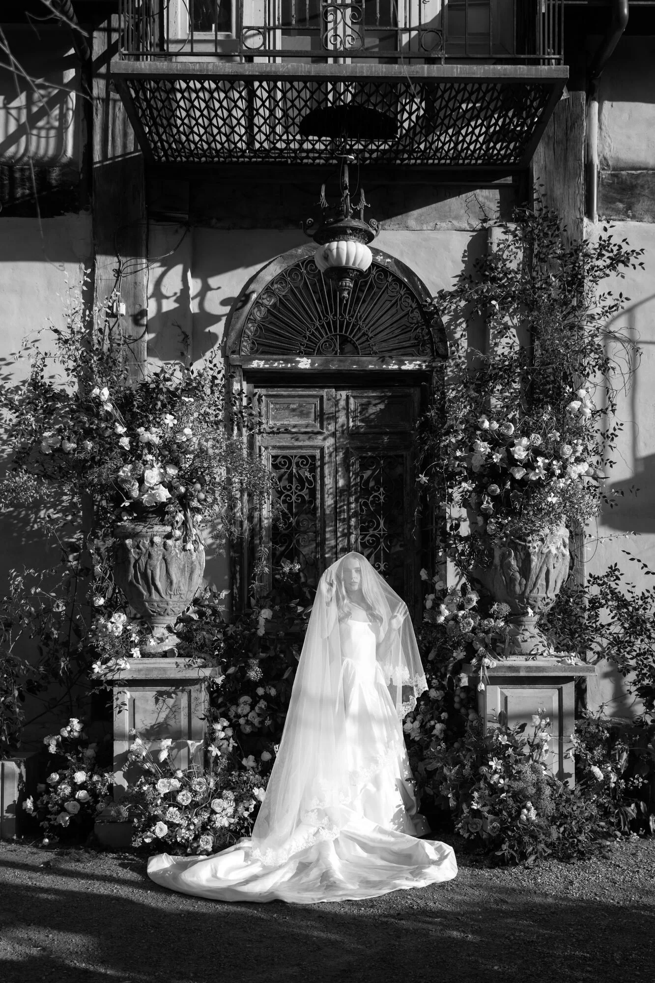 Black and white photo of a bridal mannequin wearing a wedding dress with a long train and veil, standing in front of a door adorned with flowers and greenery, with large planters on either side, and railings above.