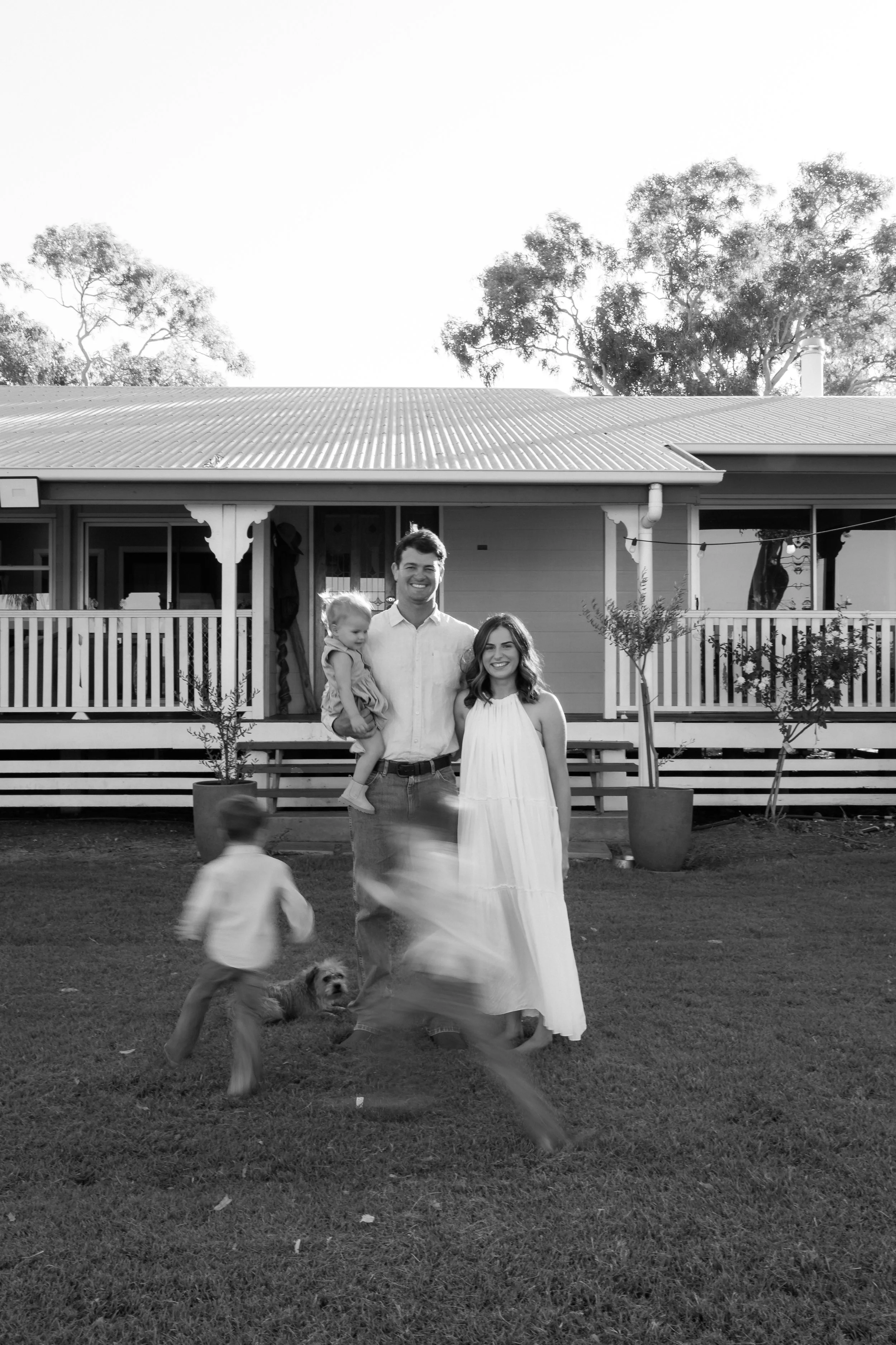 A family of five standing on lawn in front of house with porch, trees in background. Three children playing, and two adults, one woman wearing a long dress and man holding a child. Two children are blurred as they move.