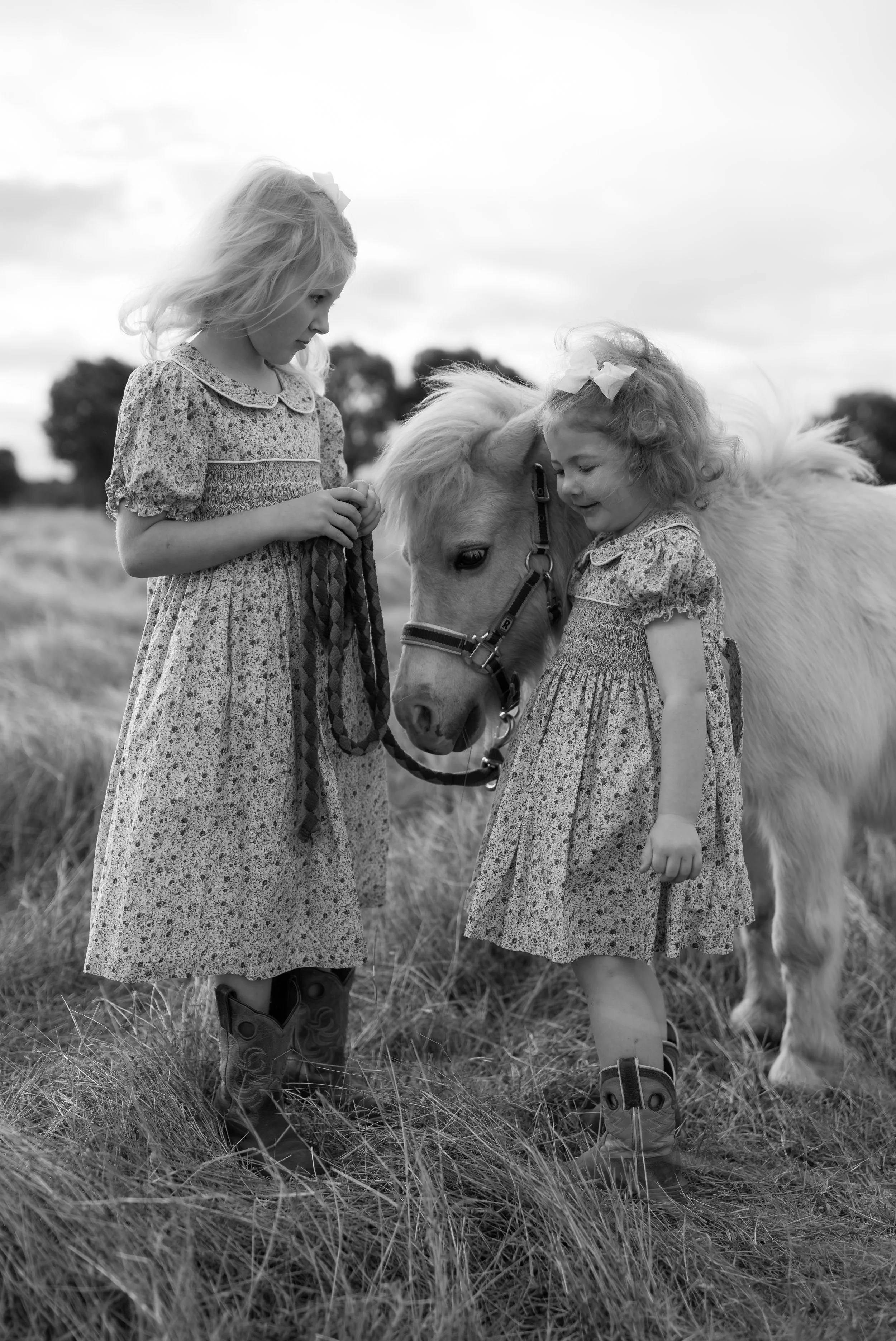 Two young girls in vintage-style dresses and cowboy boots standing on a grassy field with a small white pony, touching its head.