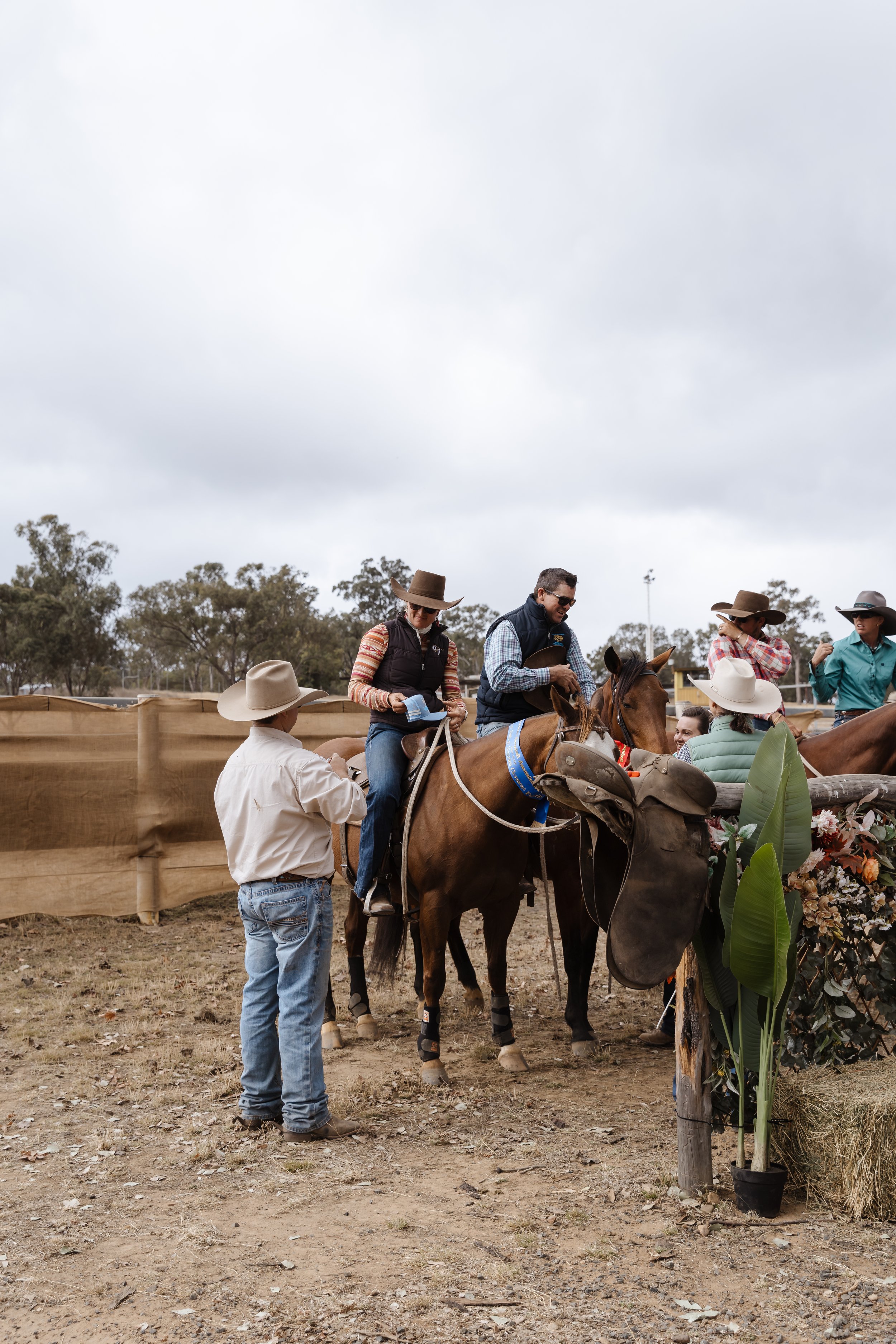 Group of people at a rodeo event, some on horseback and others standing nearby, with overcast sky and trees in the background.
