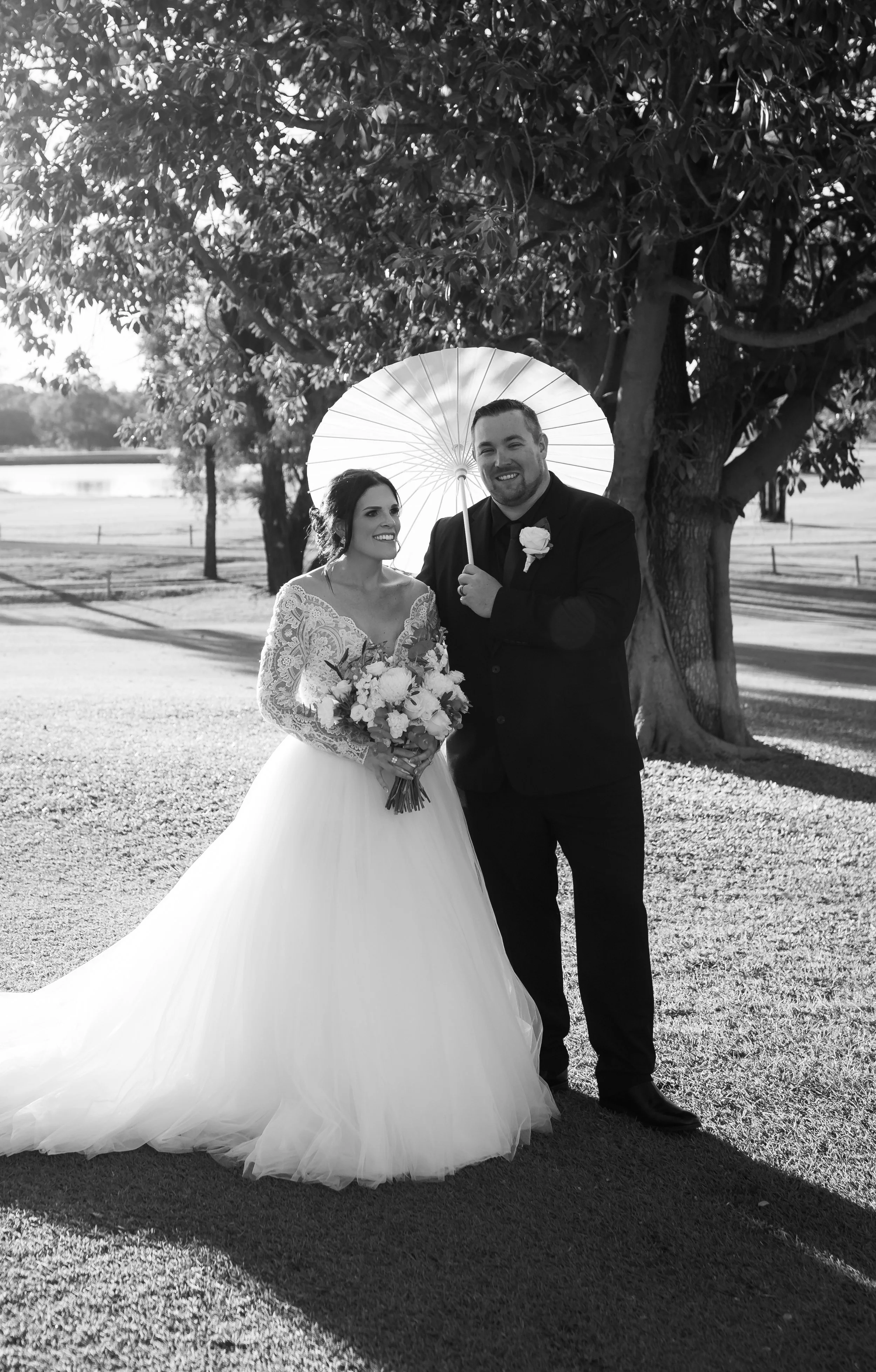 Black and white photo of a bride and groom outdoors. The bride is holding a bouquet and wearing a wedding dress with lace sleeves and a full skirt. The groom is wearing a dark suit with a boutonniere, and is holding a parasol behind him. They are sta