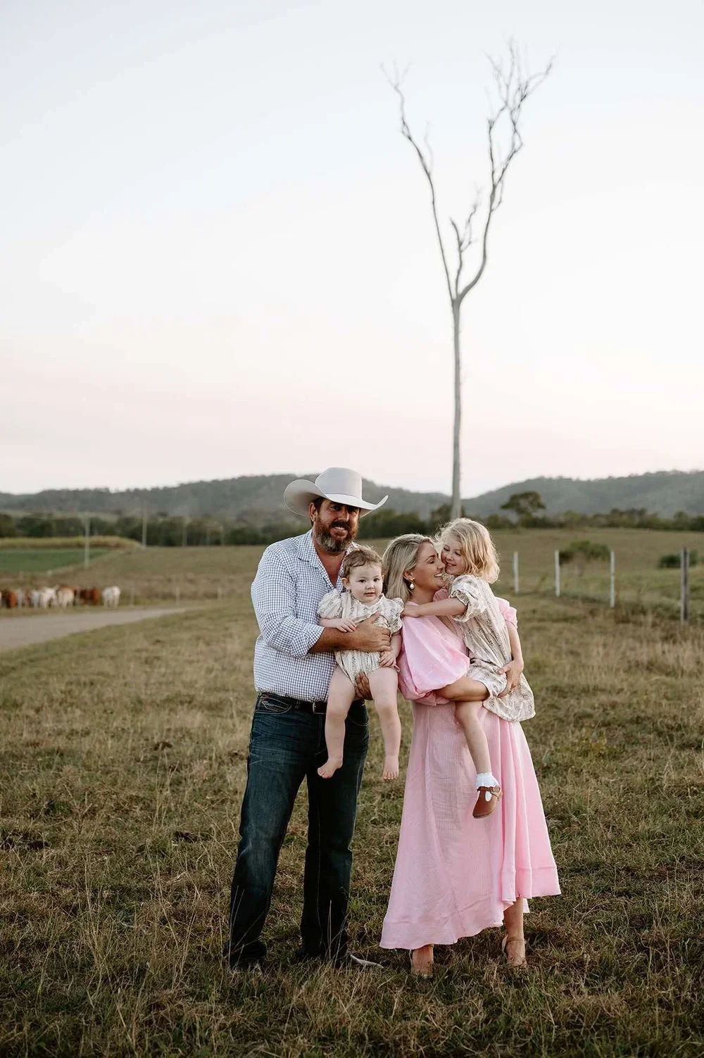 A family of four, including a man wearing a cowboy hat, a woman, and two young girls, standing on a grassy field with rolling hills and a leafless tree in the background. The man is holding one girl, and the woman is holding the other girl, both of whom are smiling and playing.