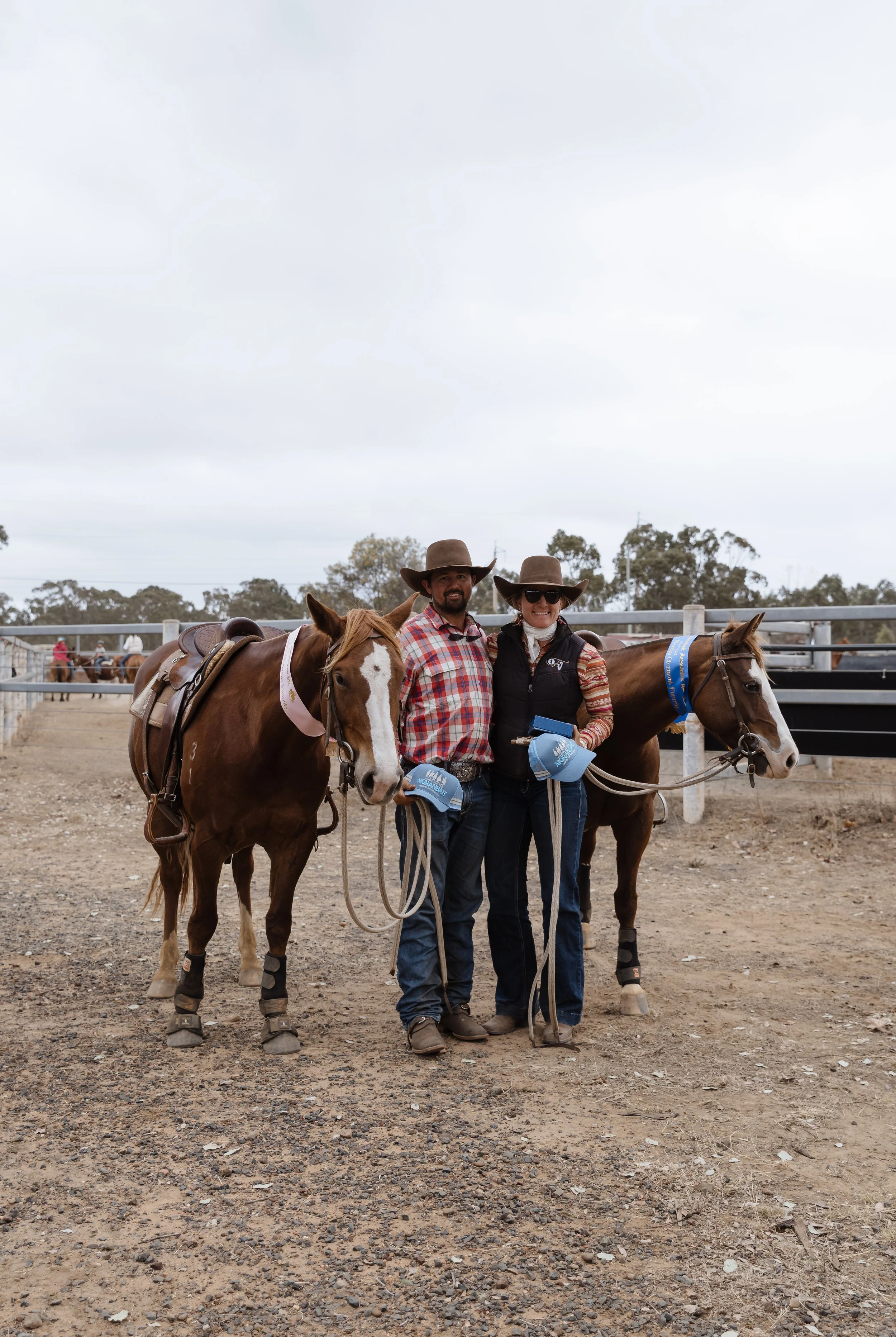 Two people standing next to two horses in an outdoor arena. Both are wearing cowboy hats. One person is smiling and holding the reins of the horse, which has a blue ribbon. The other person is also holding the reins and has a paper or booklet in hand