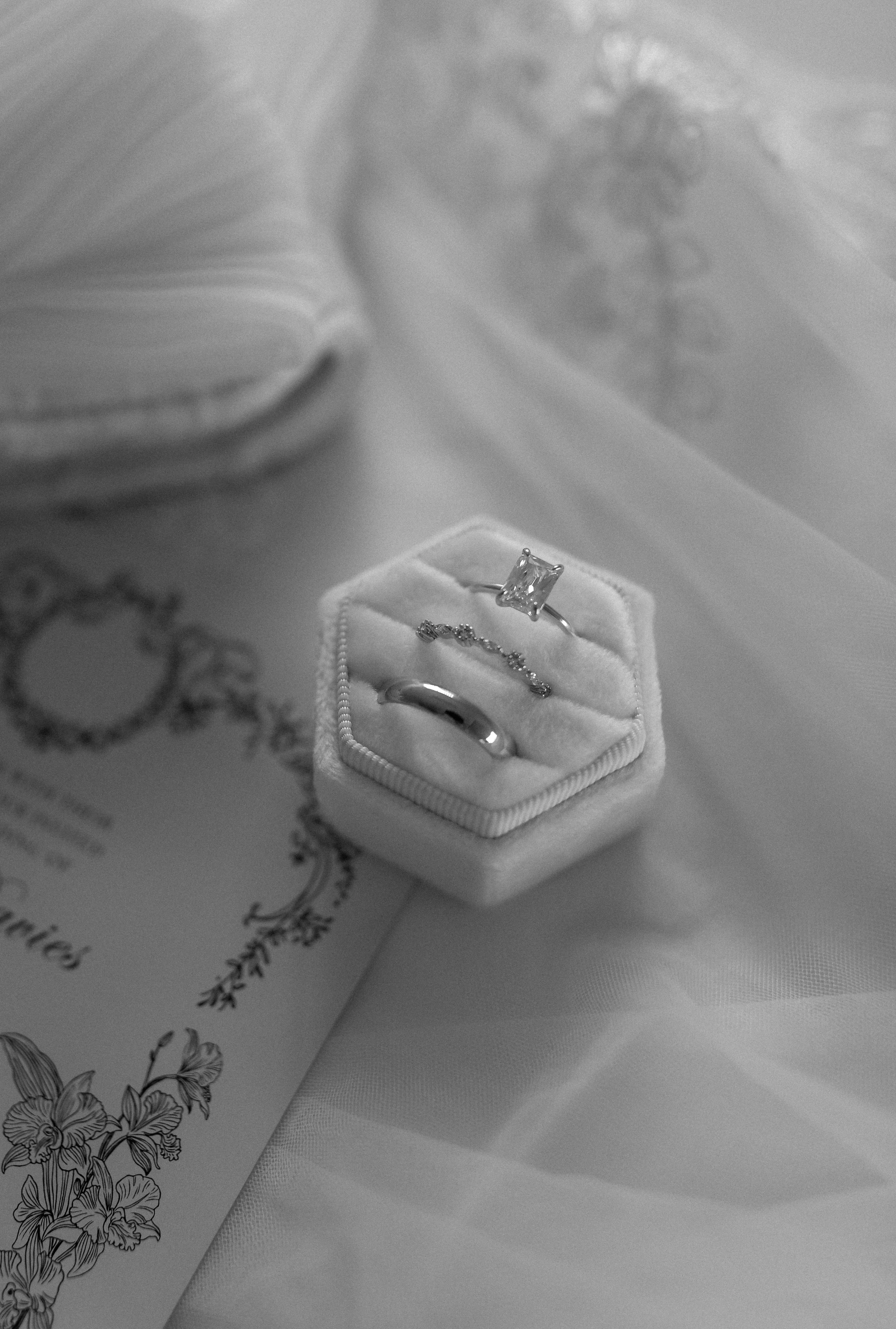 Close-up of a diamond engagement ring displayed on a velvet ring box, with a wedding band underneath, on a satin surface. In the background, a jewelry box and a card with floral illustrations are visible.