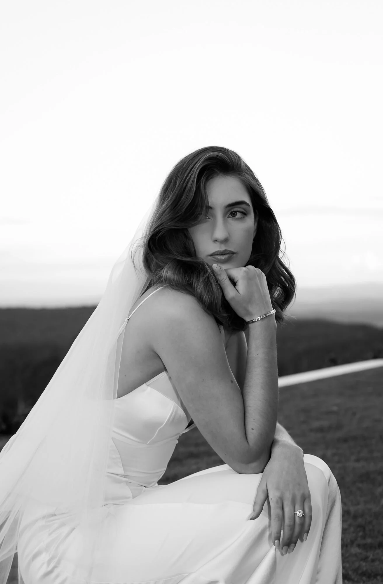 Black and white photo of a woman with long hair, wearing a white dress, sitting outdoors with a blurred landscape in the background, resting her chin on her hand, looking contemplative.