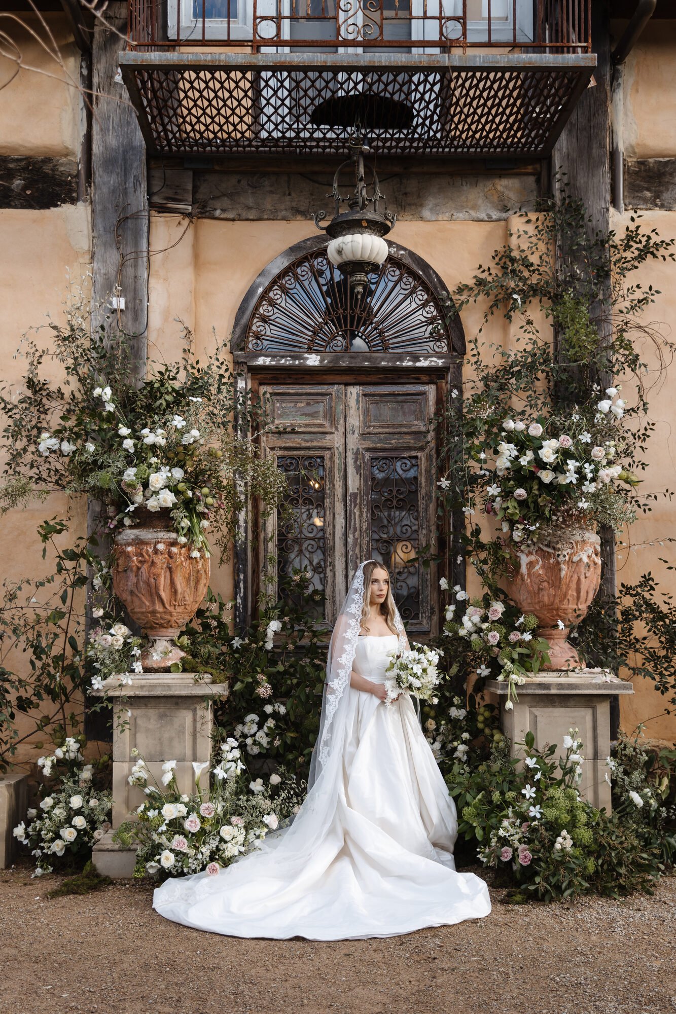A bride in a white wedding dress and veil holding a bouquet stands in front of an ornate, weathered wooden door decorated with an abundance of white and pink flowers and greenery in large urns.
