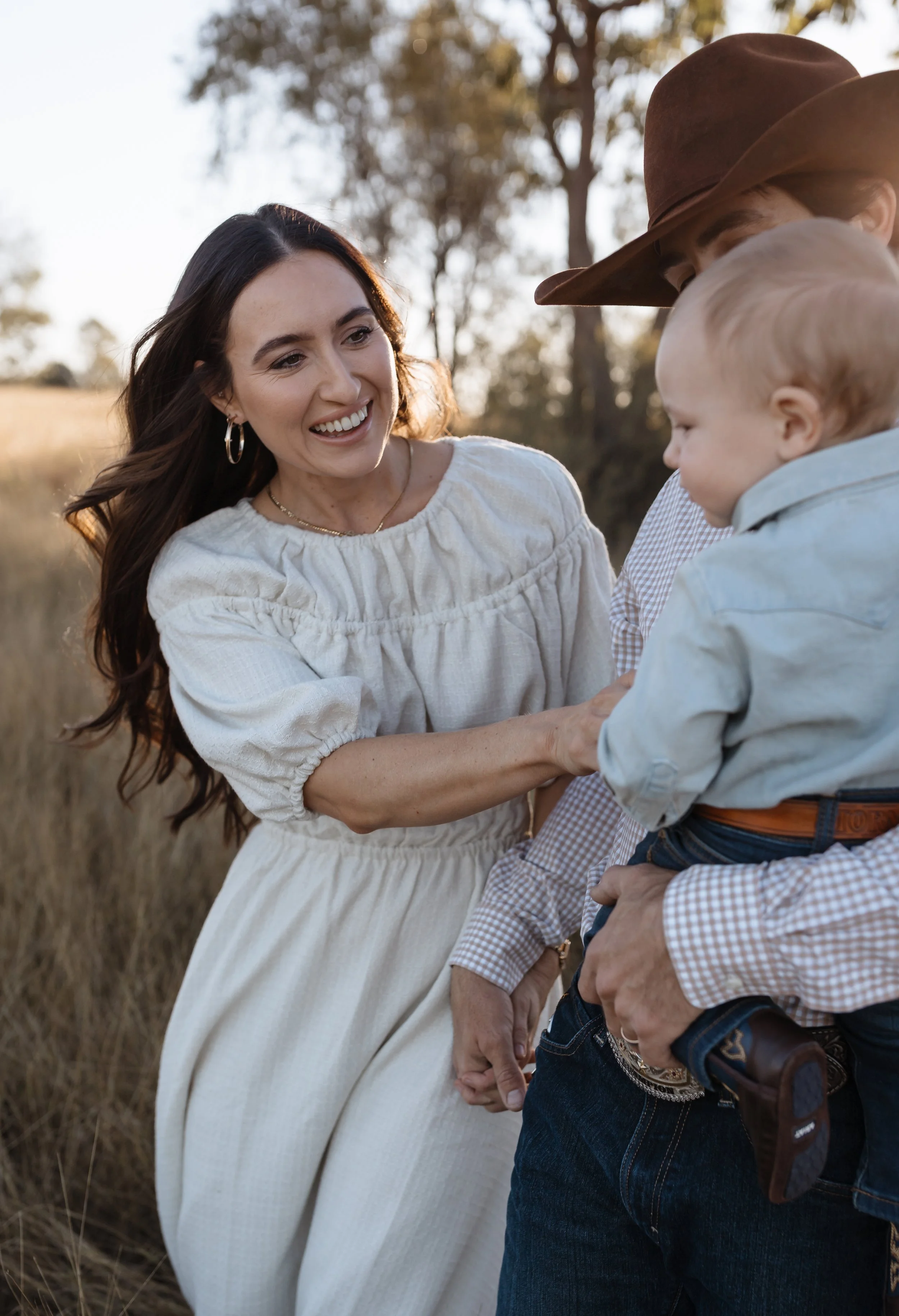 A woman dressed in a white dress smiling while holding a young boy dressed in a light blue shirt and jeans, outdoors in a park with trees in the background.