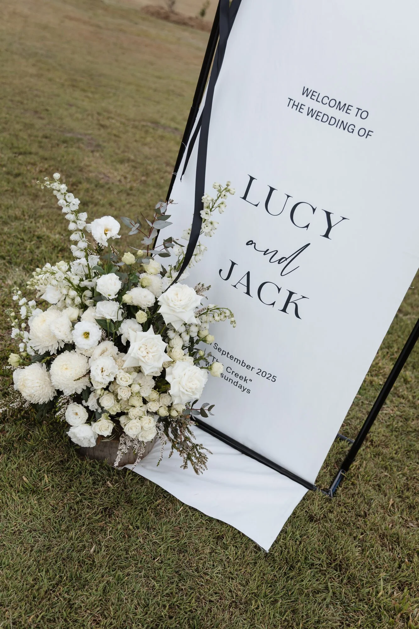 A wedding sign with white flowers and black ribbons next to it, on a grassy area.