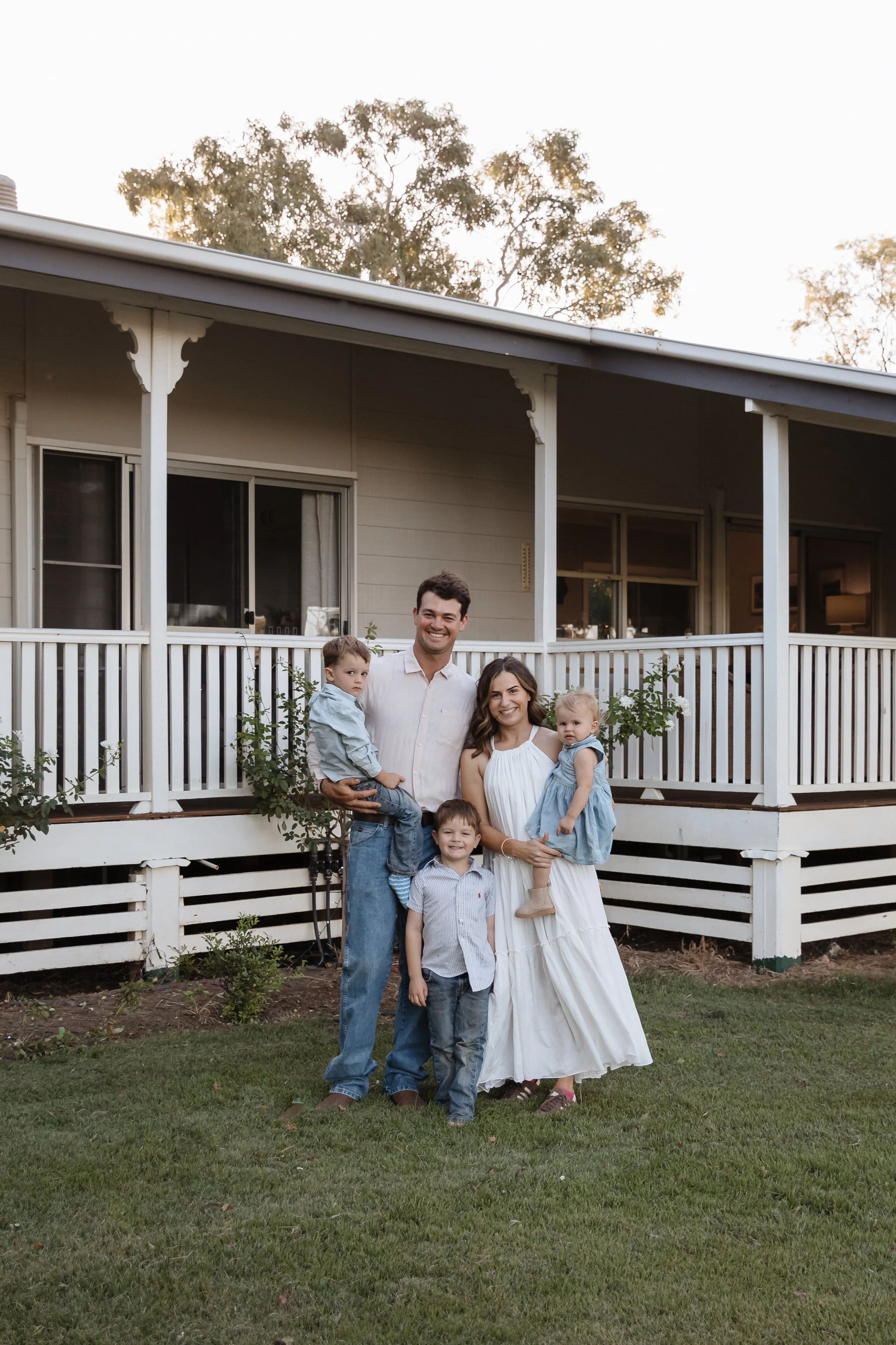Smiling family of six in front of a two-story house with a white picket fence, trees in the background, and a grassy yard, during daytime.