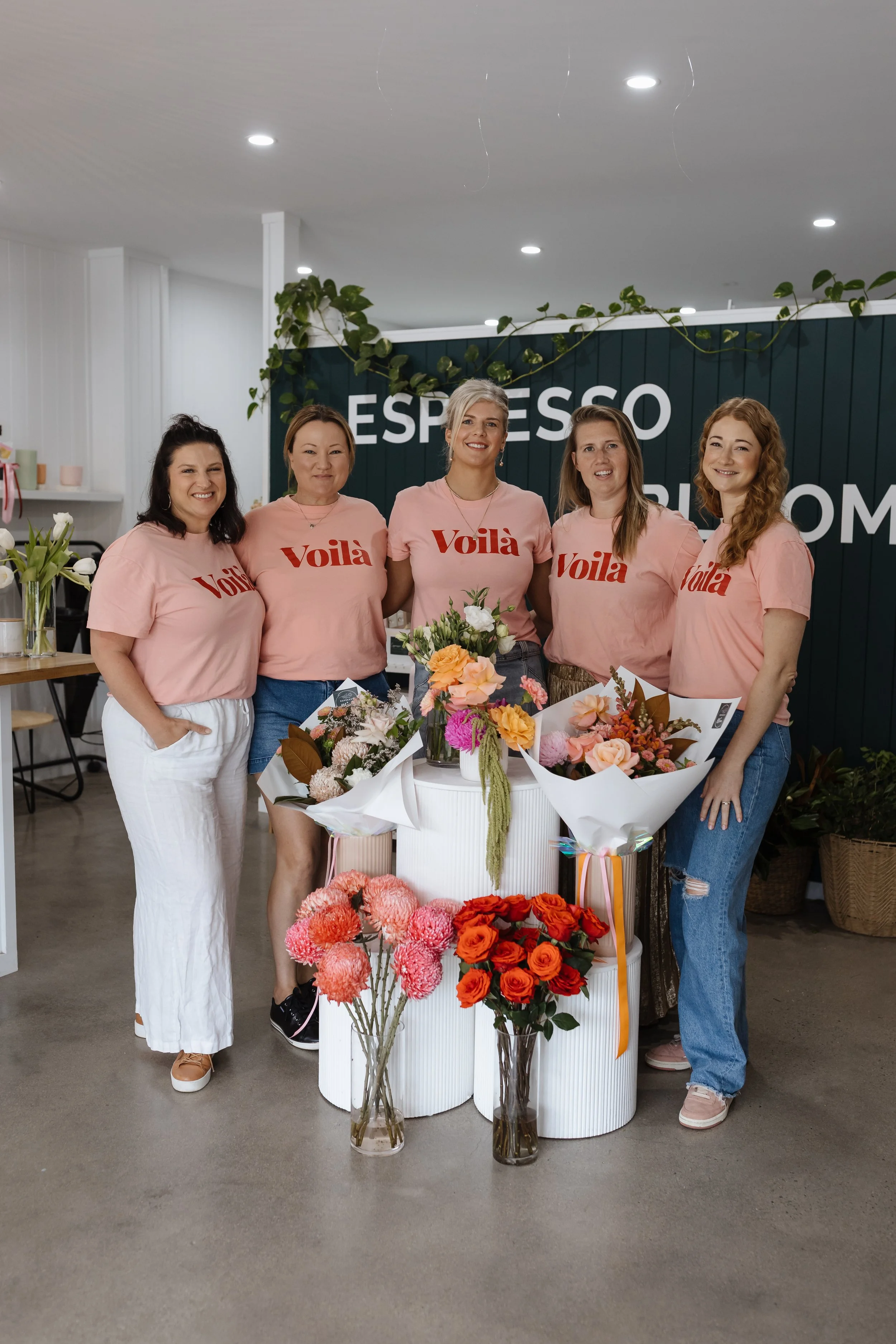 Five women wearing matching pink shirts with the word "Voilà" standing behind a table with bouquets of flowers. The background has a dark green sign with part of the word "Espresso" visible.