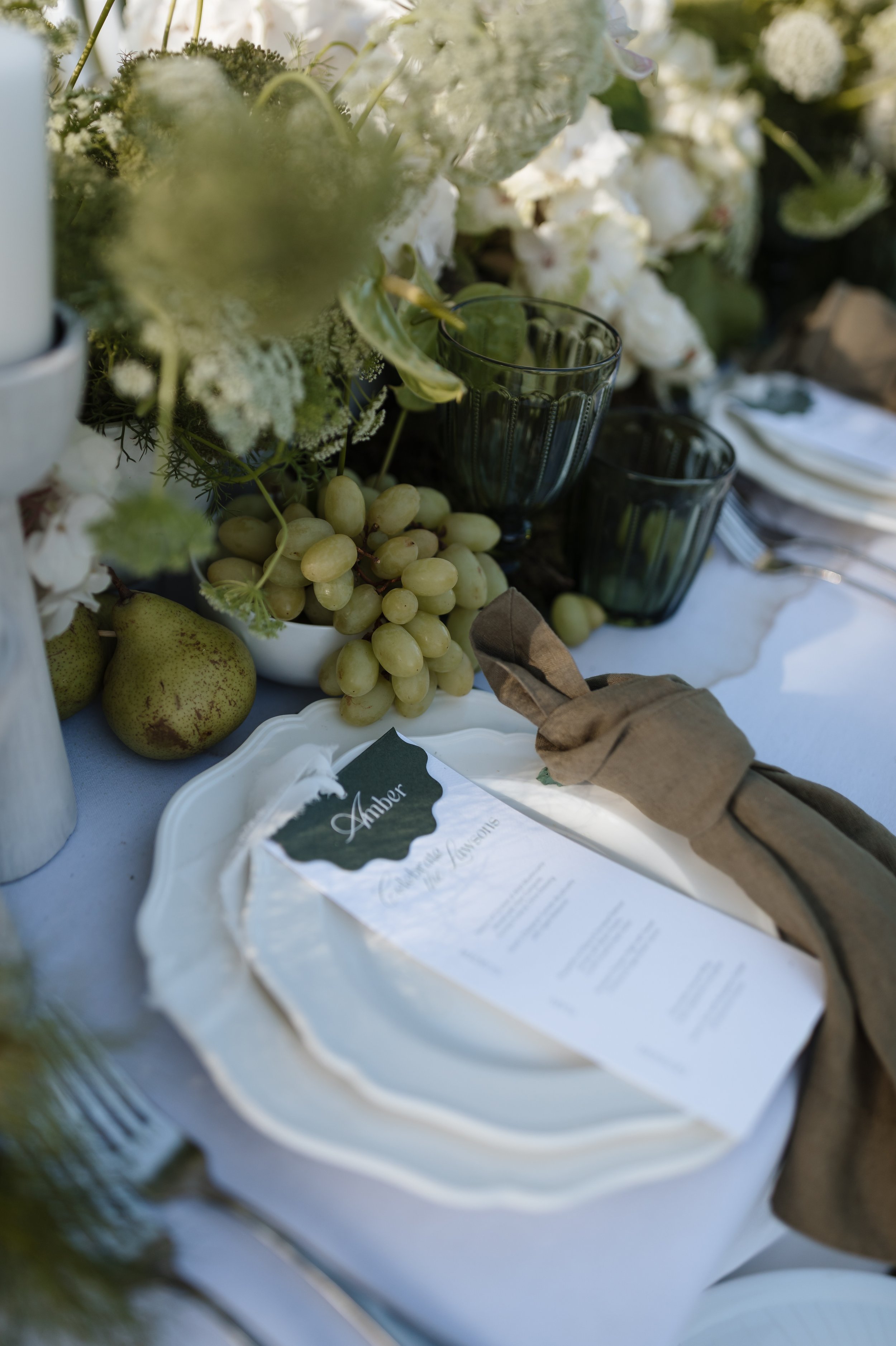 A table with a floral centerpiece, grapevines, and pears, along with in focus glasses and a menu labeled 'Amber'.