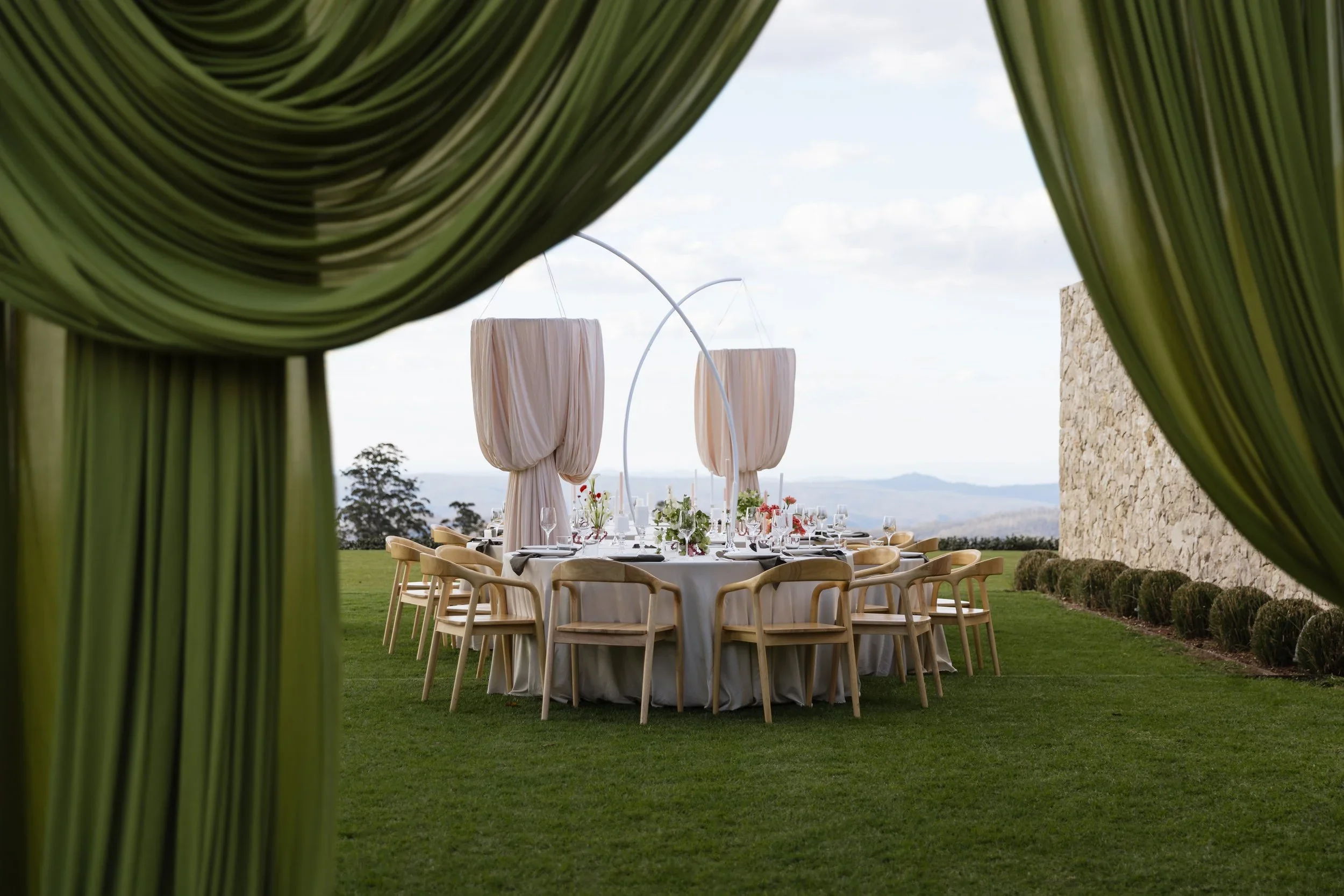 Elegant outdoor dining setup with a round table, white tablecloth, and chairs, seen through green draped curtains, with scenic landscape in the background.