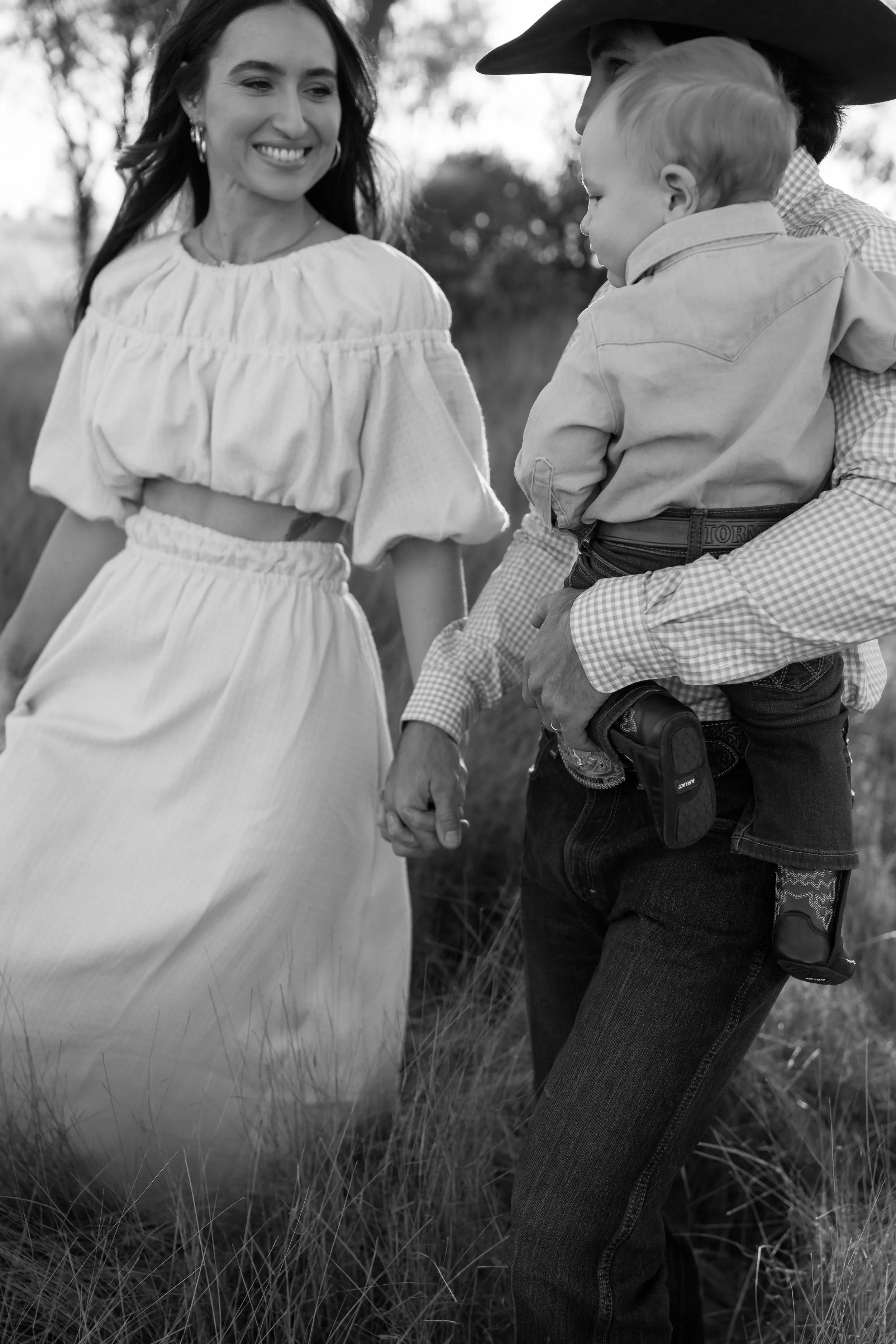 A woman and a man holding a young boy by a field, smiling and looking at each other in black and white. The woman is wearing a ruffled, off-the-shoulder dress, and the man is wearing a hat and checkered shirt.