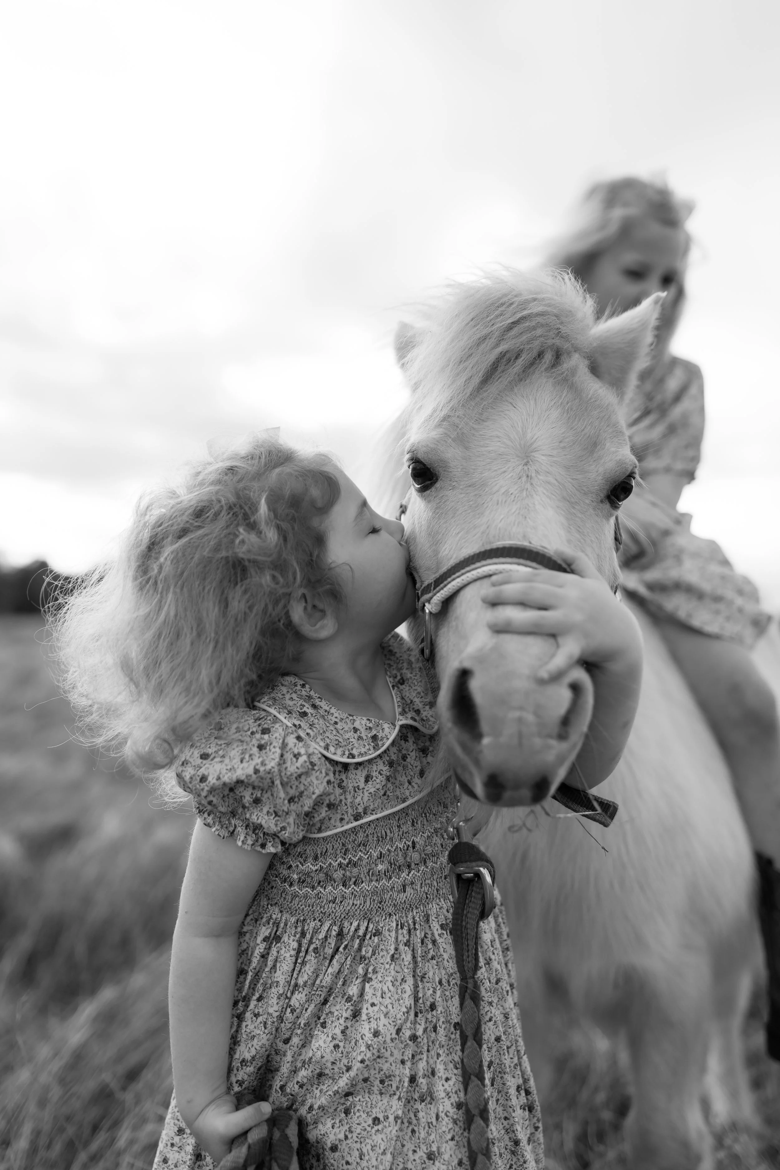 A young girl with curly hair kisses a white horse on the nose as another girl, partially visible, stands nearby. The scene is outdoors on a grassy field with a cloudy sky.