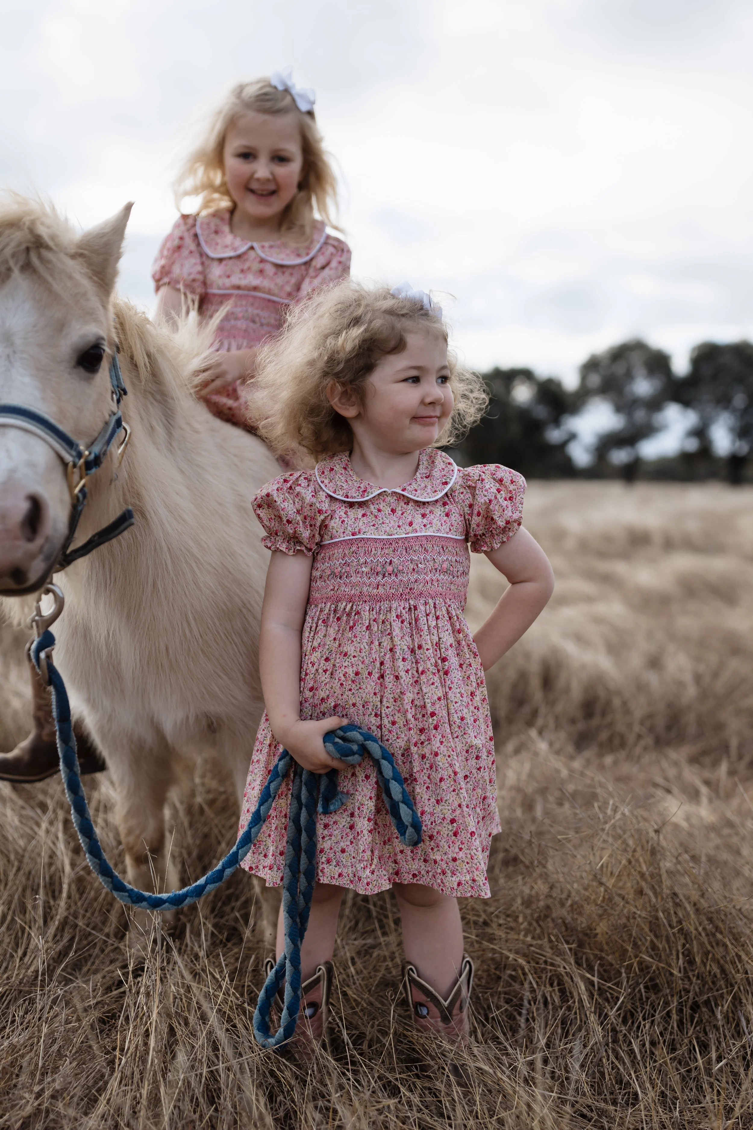 Two young girls with curly hair and floral dresses standing in a field with a white horse, holding its blue rope, under a cloudy sky.