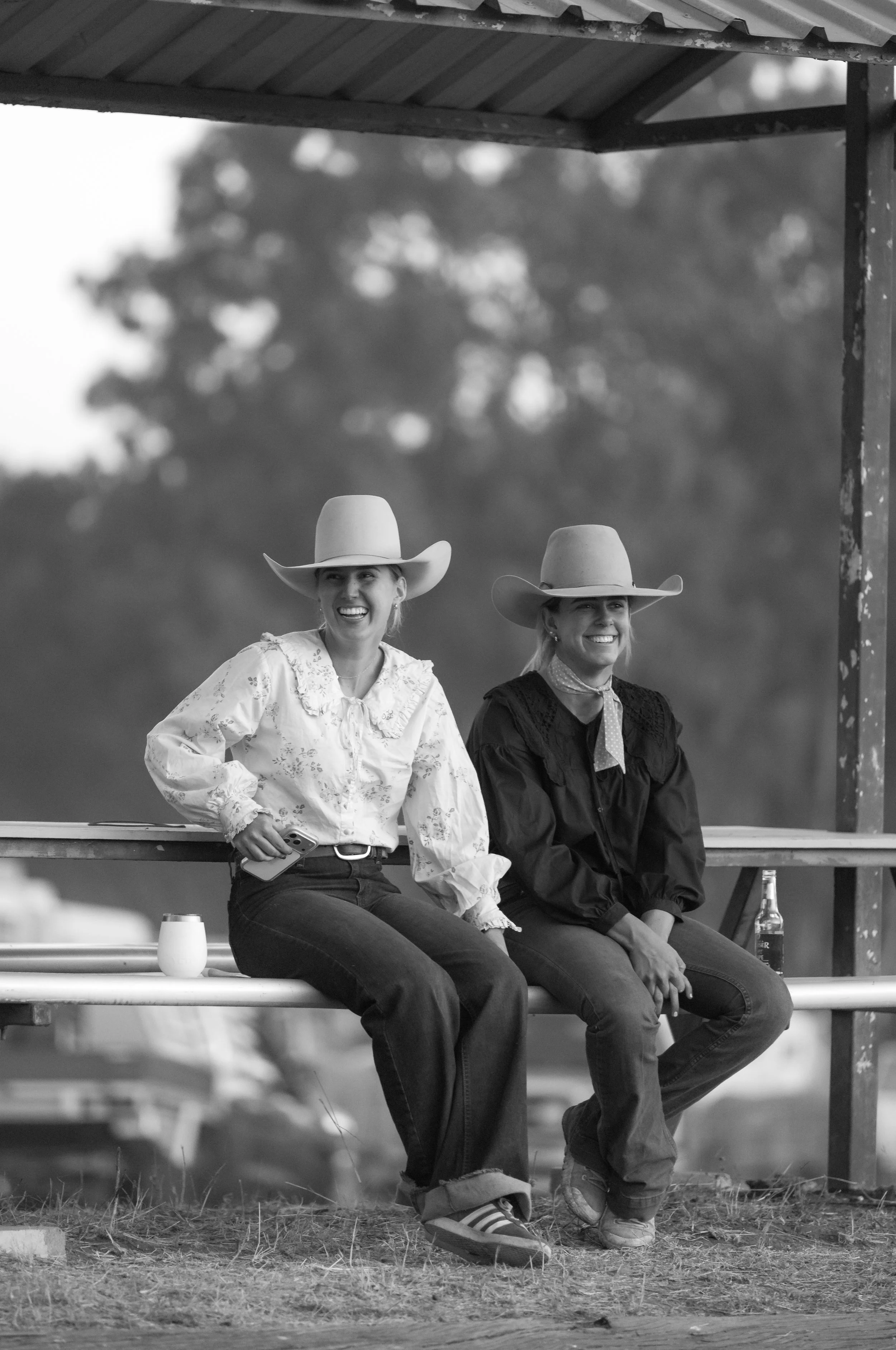 Two women wearing cowboy hats sit on a bench at an outdoor event, smiling and laughing. One woman holds a smartphone, and there are drinks on the bench in front of them. The background shows trees and an overcast sky.