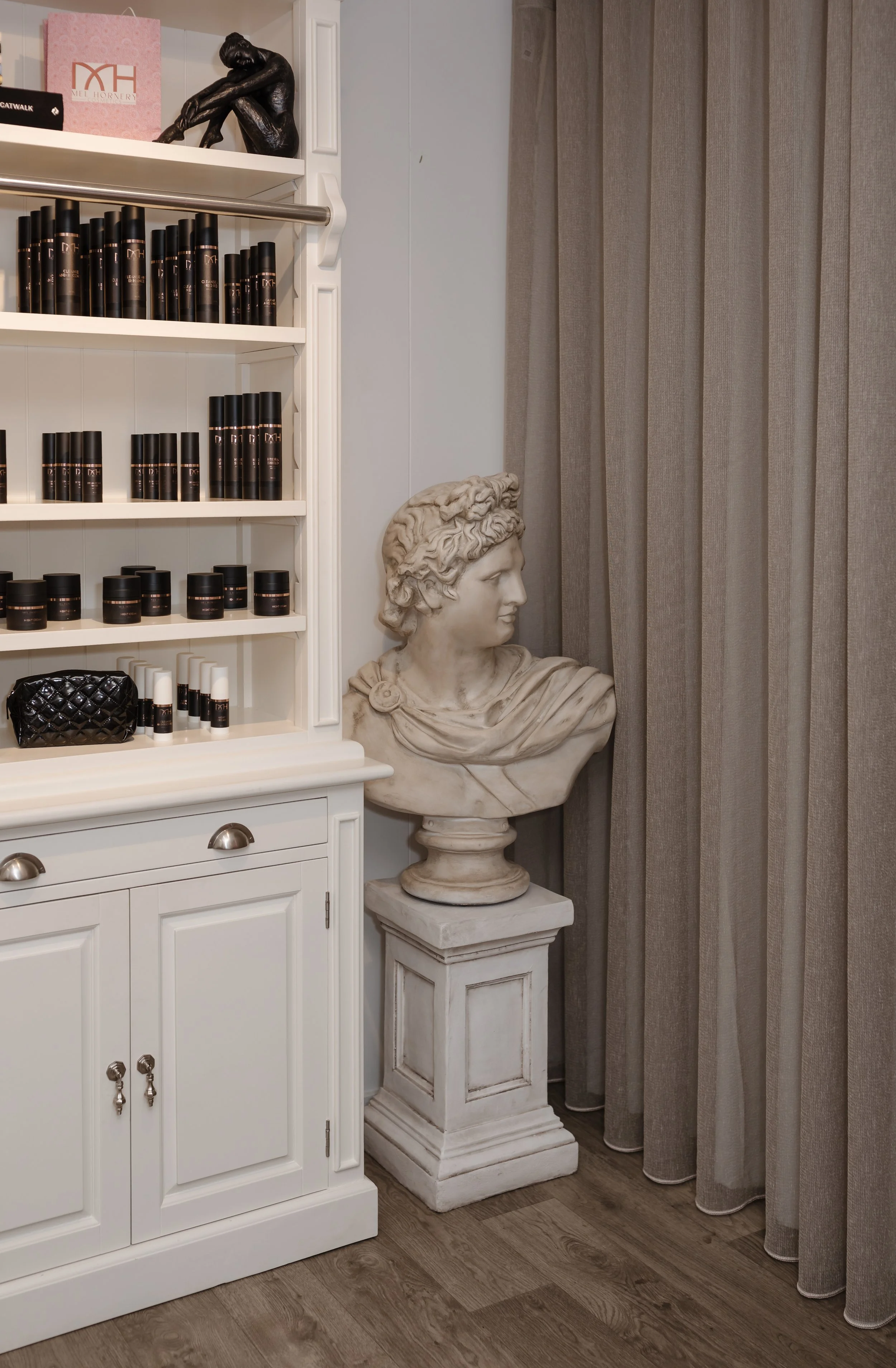 A white cabinet with shelves filled with black bottles, a small black quilted purse, and some white items. Next to the cabinet is a classical-style bust sculpture of a person, placed on a white pedestal and surrounded by beige curtains.