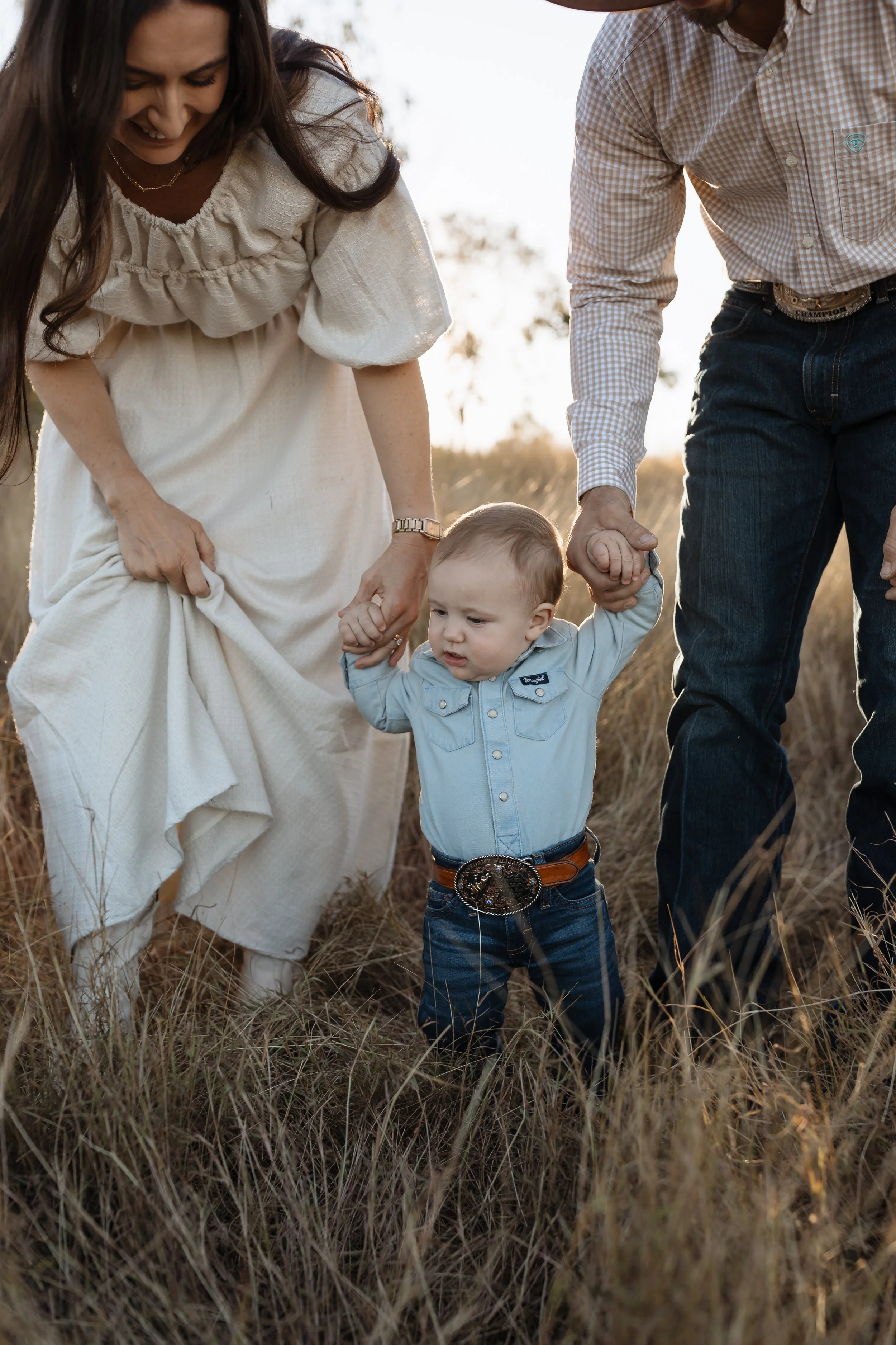 A family, including a woman, a man, and a young boy, holding hands and walking through a grassy field during sunset.