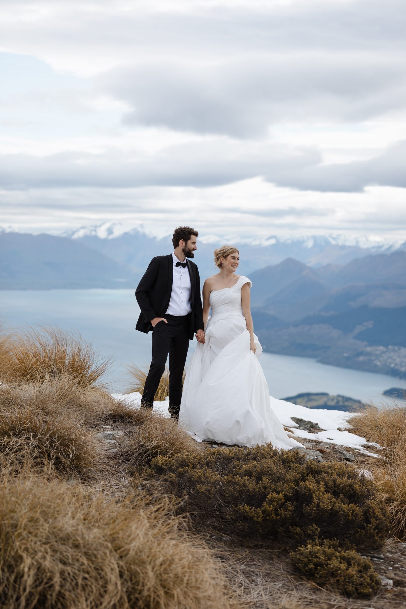 A bride and groom holding hands and walking outdoors in a mountainous landscape with water, snow, and clouds in the background.