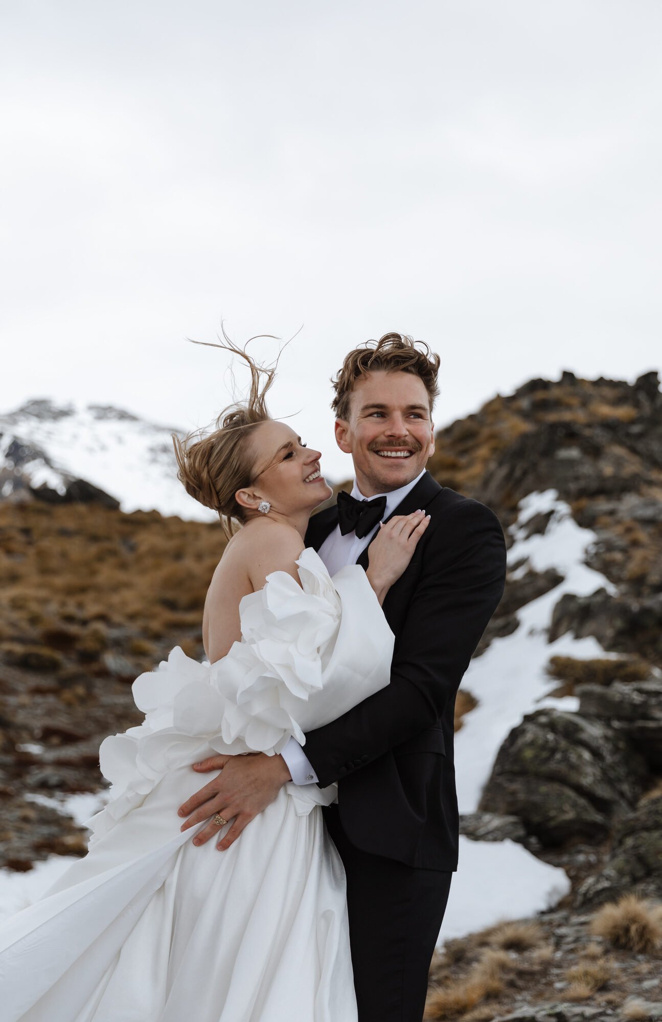 A smiling man in a black tuxedo holding a woman in a white wedding dress outdoors in a mountainous area with snow.