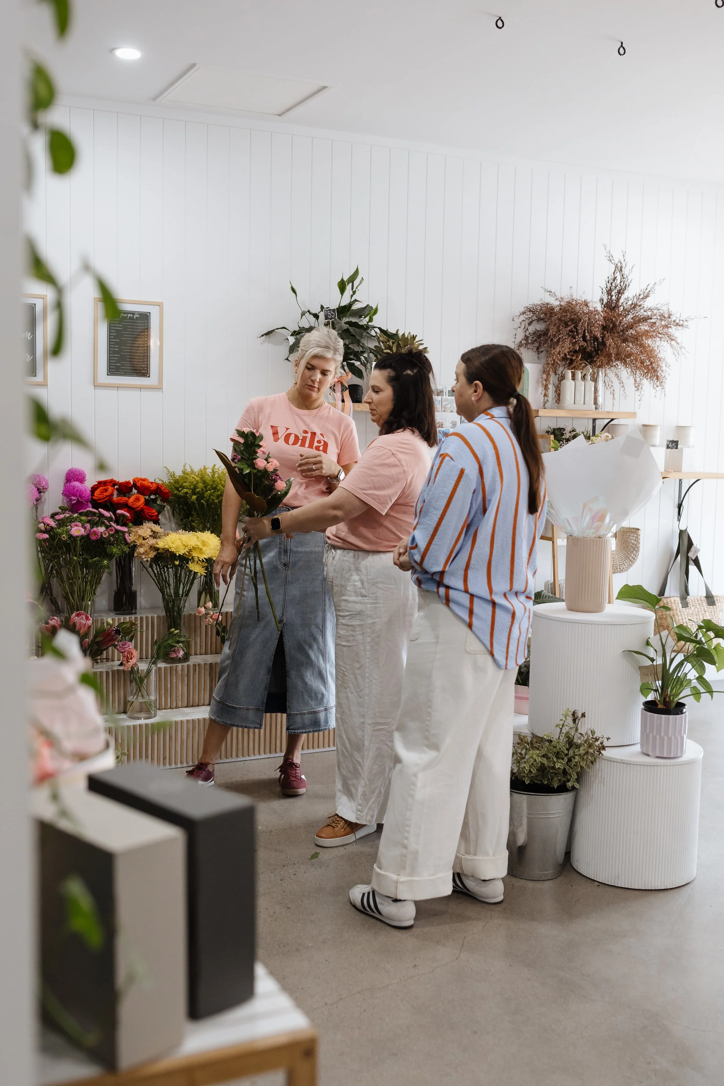 Three women inside a flower shop looking at a bouquet of pink roses. The shop is decorated with various flowers and potted plants, with white walls and wooden shelves.