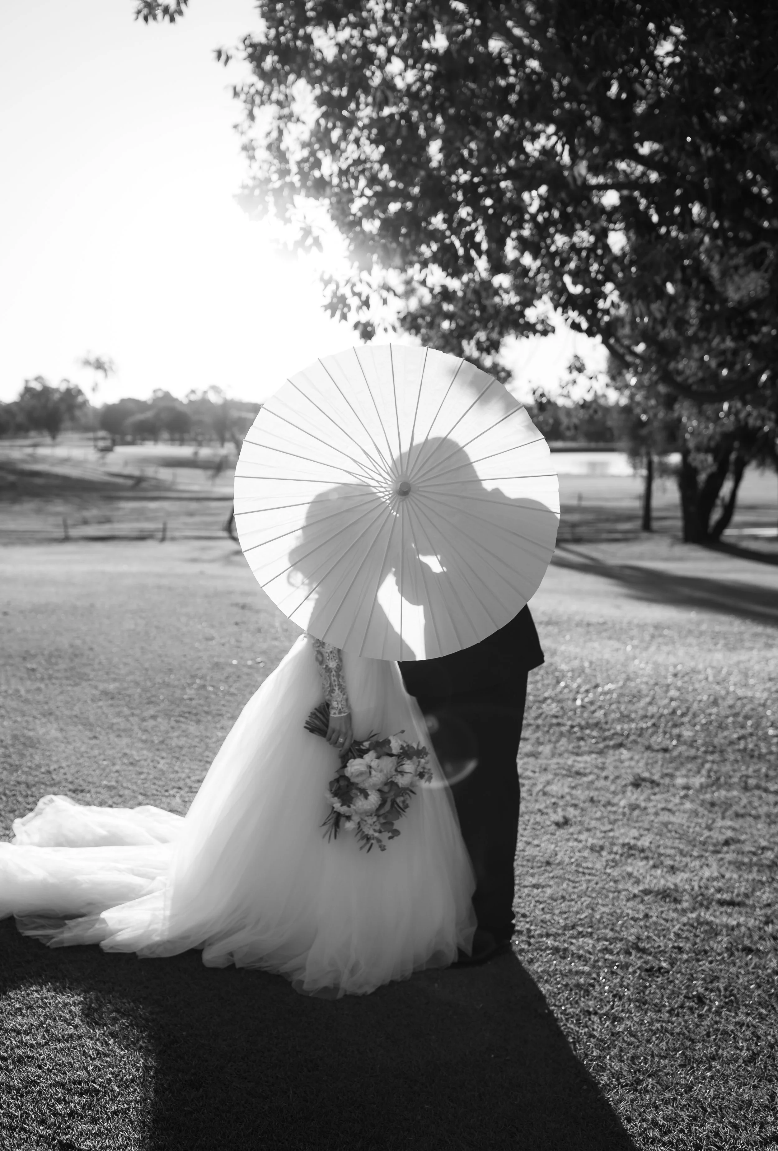 A bride and groom standing outdoors with a large sun behind them, holding a parasol that obscures their faces. The bride wears a white wedding gown and the groom is dressed in a dark suit. The scene is in a park with trees and a body of water in the 