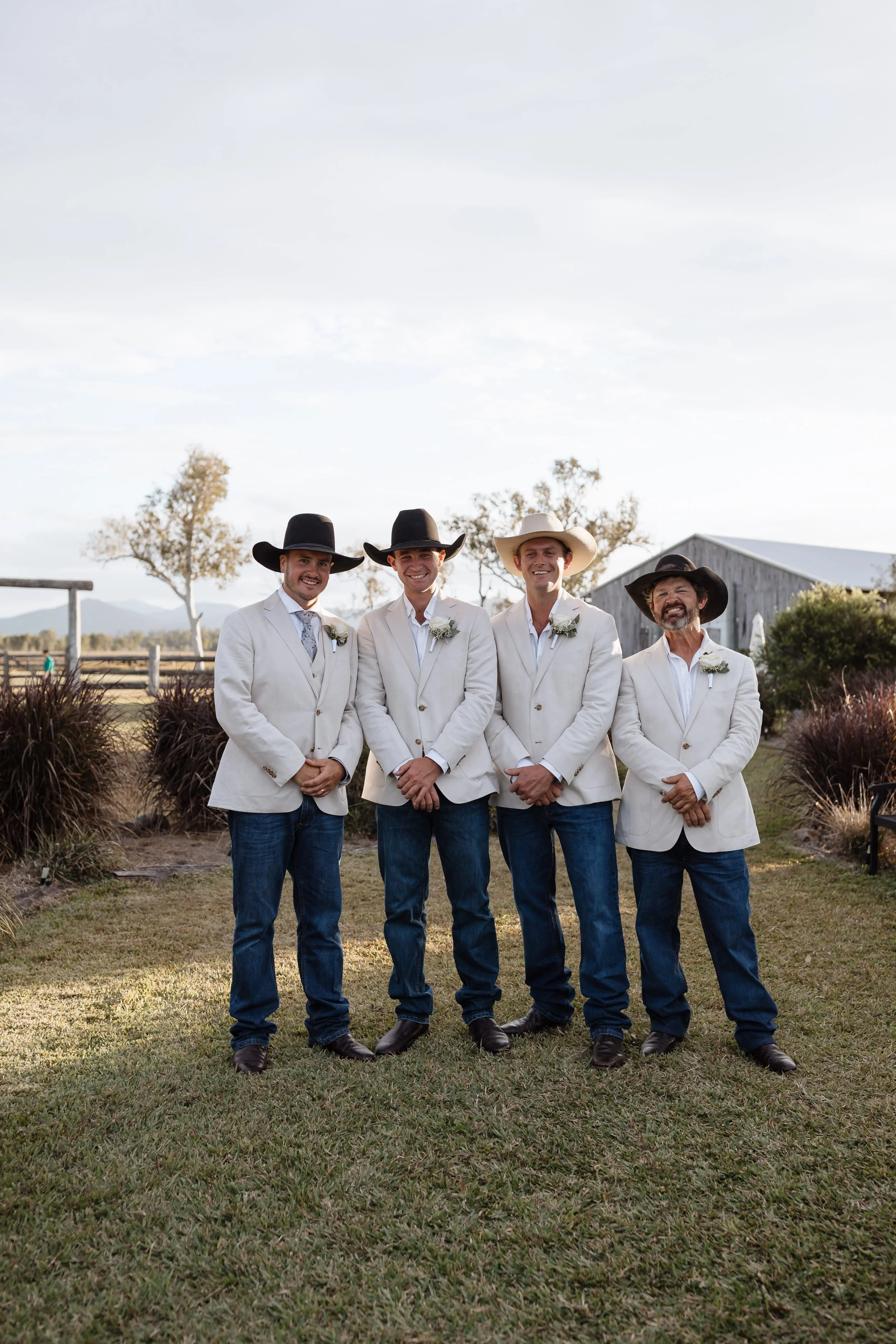 Four men standing outdoors on grass in matching white blazers, jeans, and boots, wearing cowboy hats, with a barn and trees in the background, smiling for a photo.