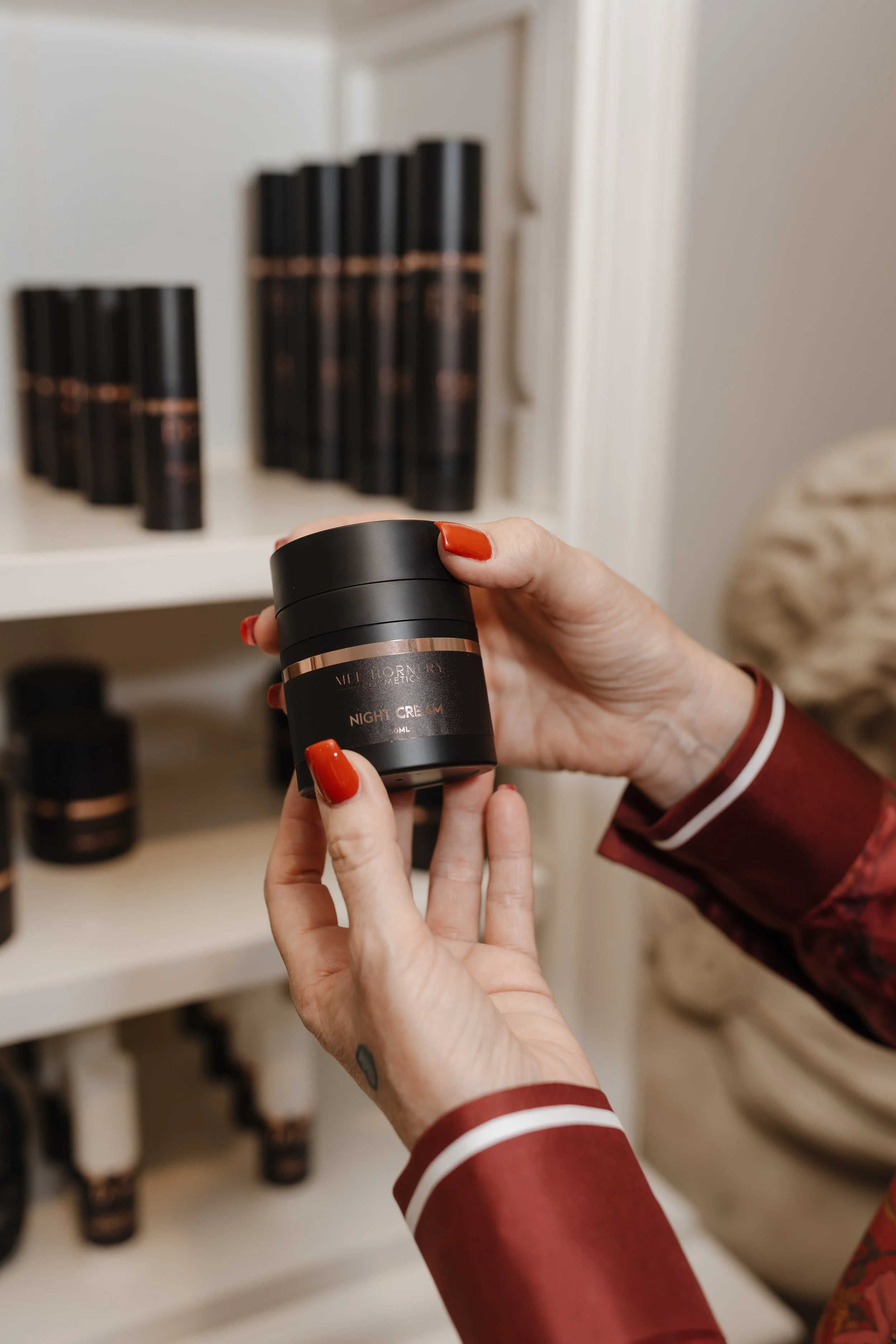 Person with red painted nails holding a container of night cream in front of a shelf filled with similar containers.