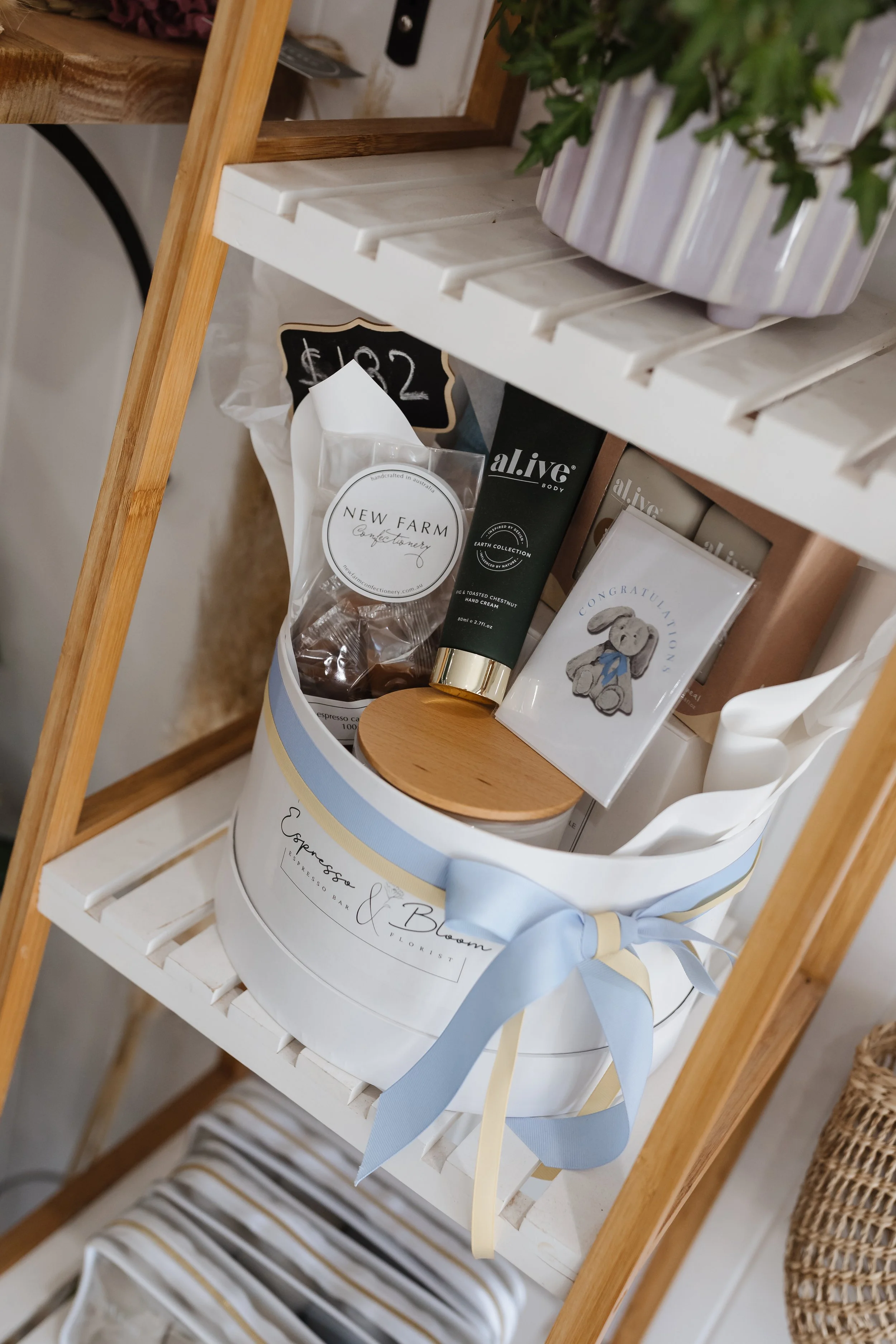 A white shopping basket with gold and light blue ribbon, holding a hand cream, greeting card, and other items, placed on a white wooden shelf in a cozy home setting.