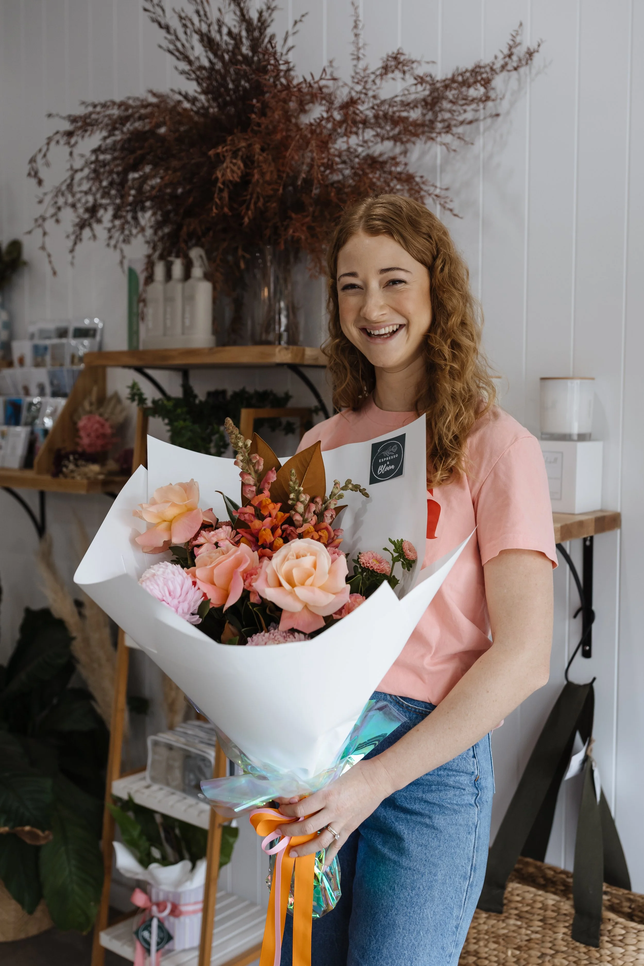 A woman with curly red hair smiling and holding a bouquet of pink and peach flowers wrapped in white paper, in a flower shop.
