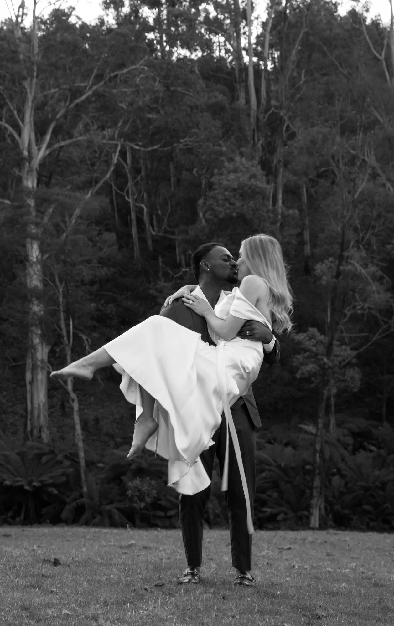 A man in a suit carrying a woman in a white dress outdoors with trees in the background, in black and white