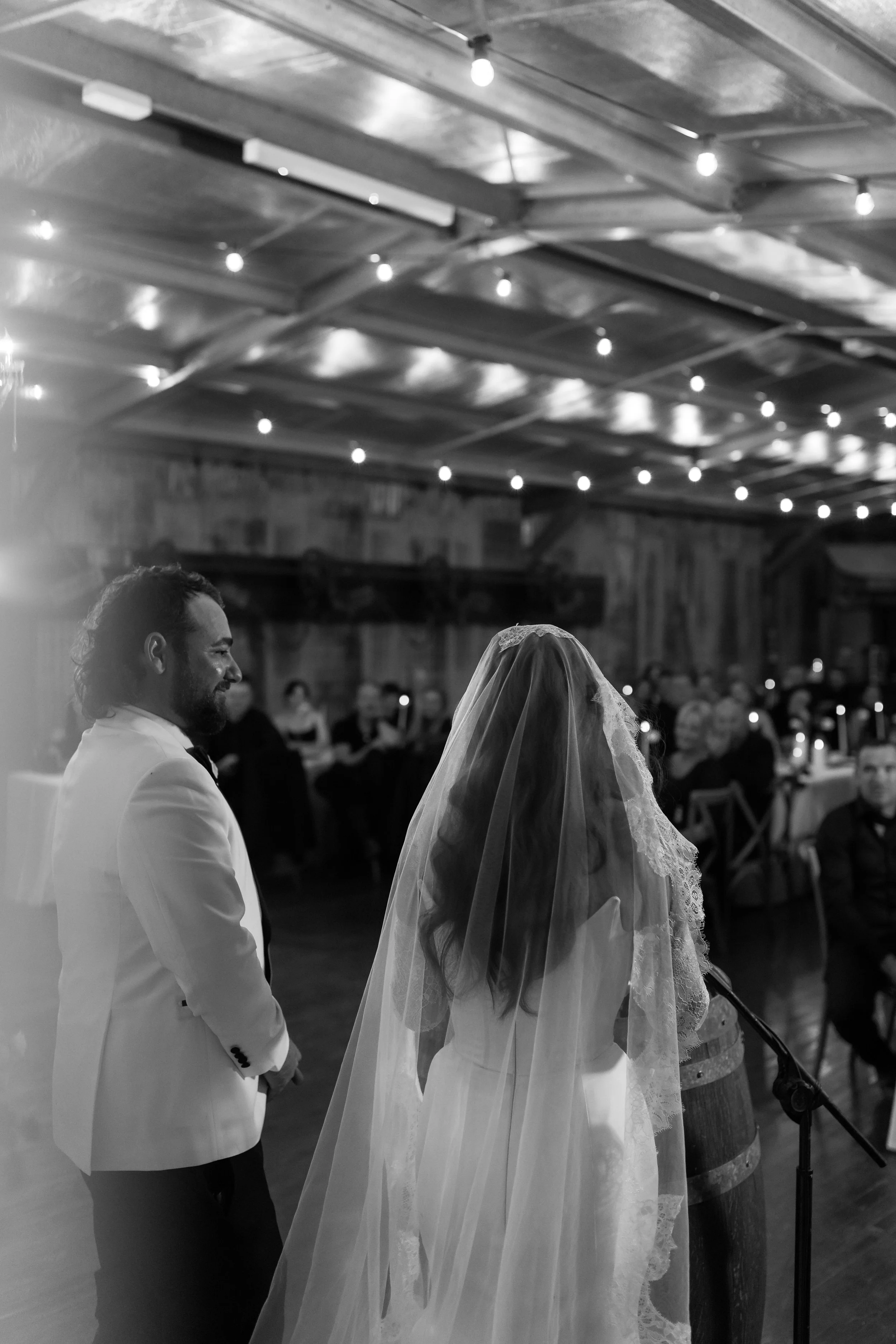 Black and white photo of a wedding ceremony with a man in a light-colored suit and a woman in a wedding dress with a veil, standing in front of a crowd inside a rustic venue decorated with string lights.