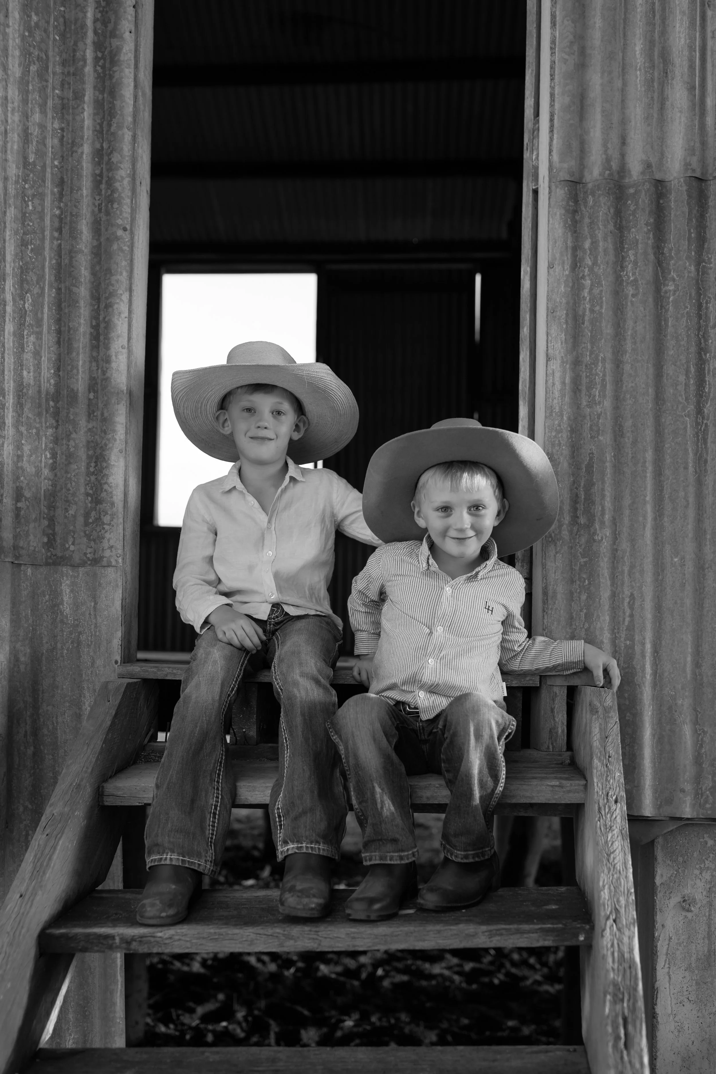 Two young boys with large hats sitting on wooden stairs outside a barn or shed, in black and white.