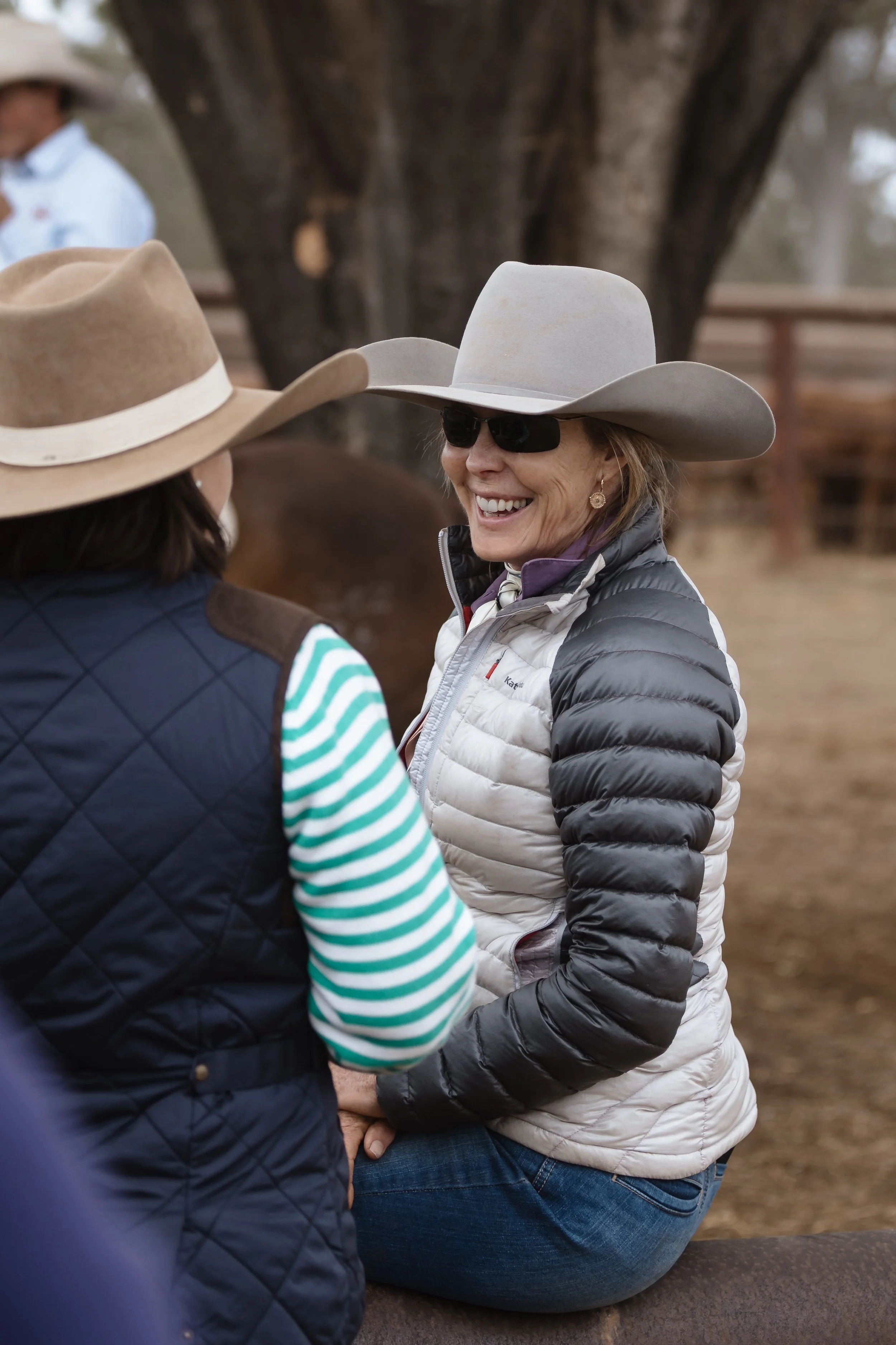 A woman with a gray cowboy hat and sunglasses smiling while talking to another person with a beige cowboy hat in an outdoor setting with a large tree in the background.