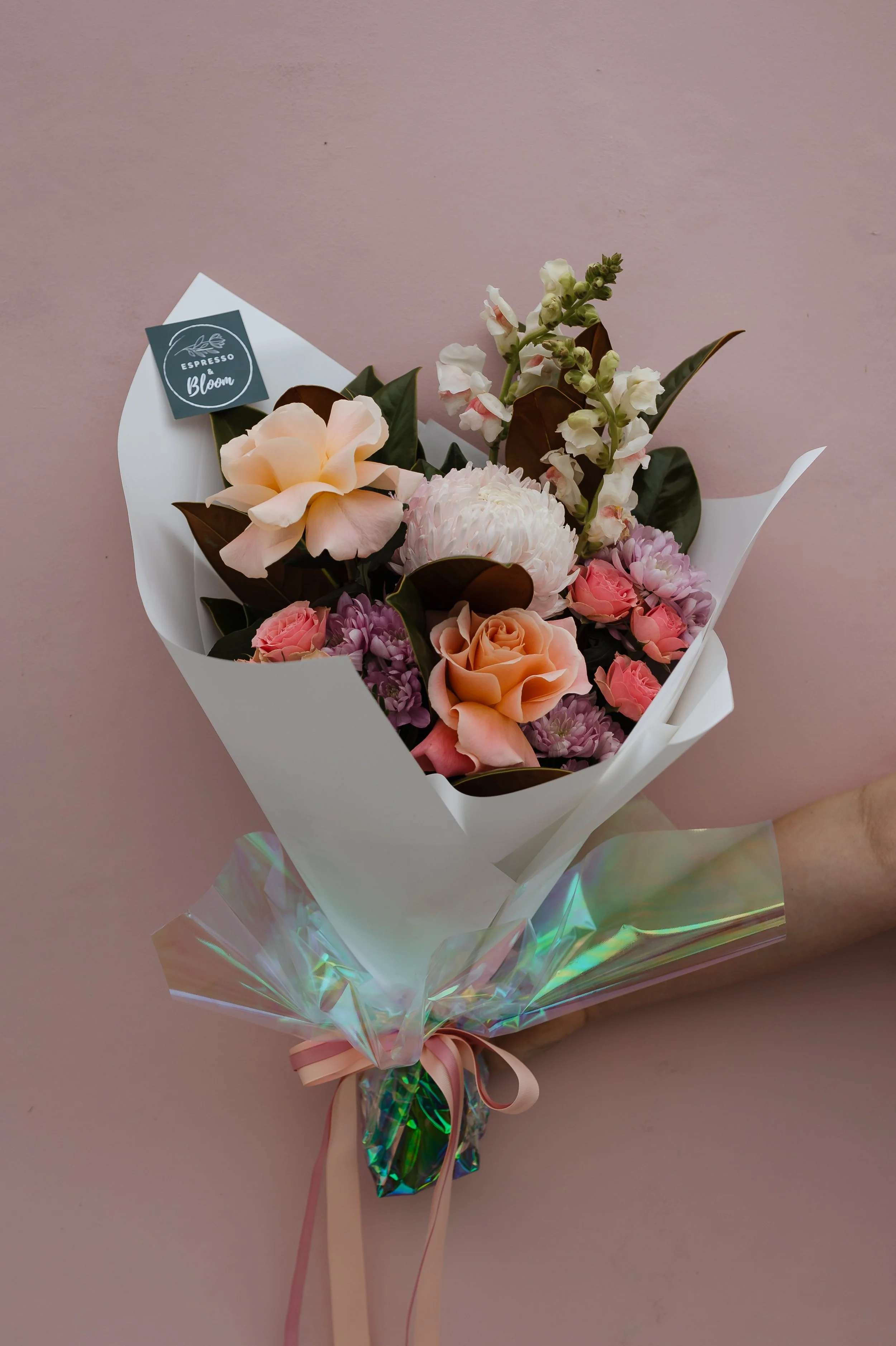Bouquet of pink, peach, and white flowers wrapped in white paper with iridescent cellophane and a pink ribbon, against a pink wall.