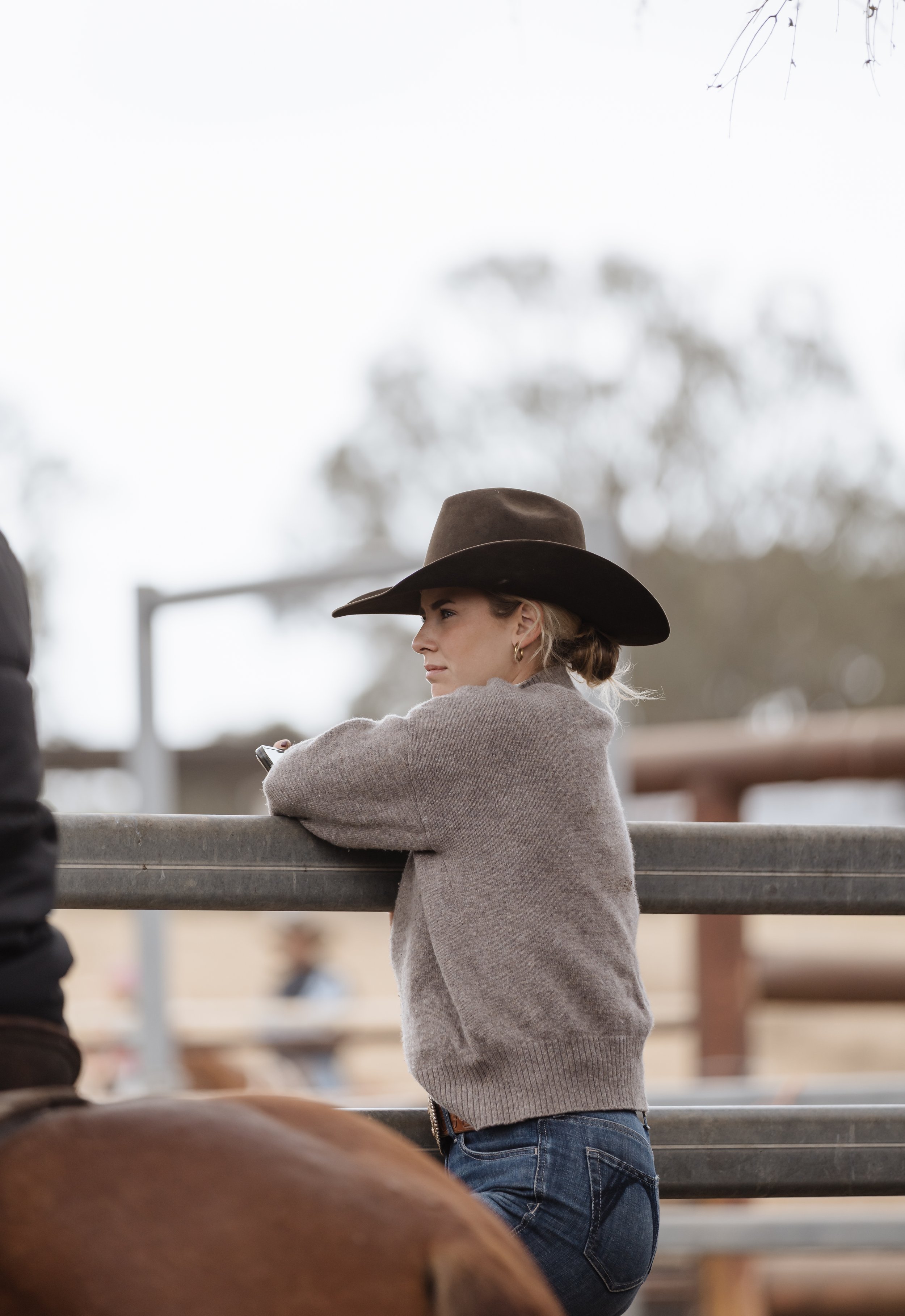 A woman wearing a brown cowboy hat, hoop earrings, a gray sweater, and jeans, leaning on a rail at an outdoor ranch with trees and a cloudy sky in the background.