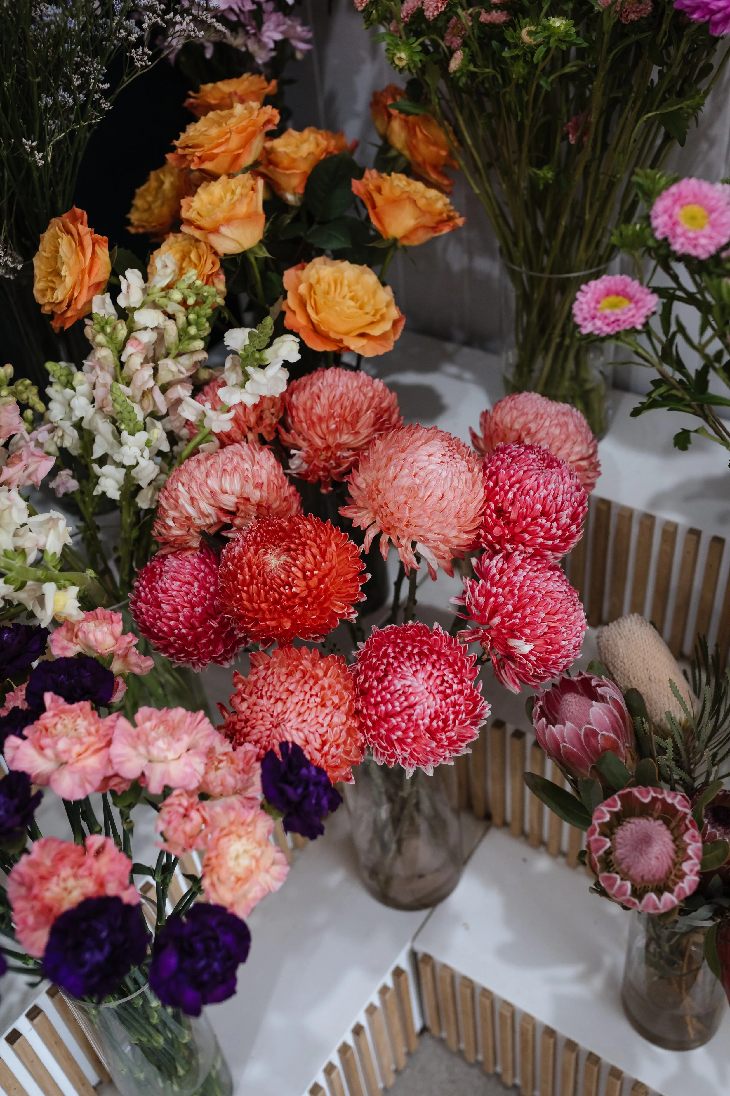 Various colorful flowers in glass vases on display, including peach roses, pink chrysanthemums, white snapdragons, and purple lisianthus, arranged on a white and wooden table.
