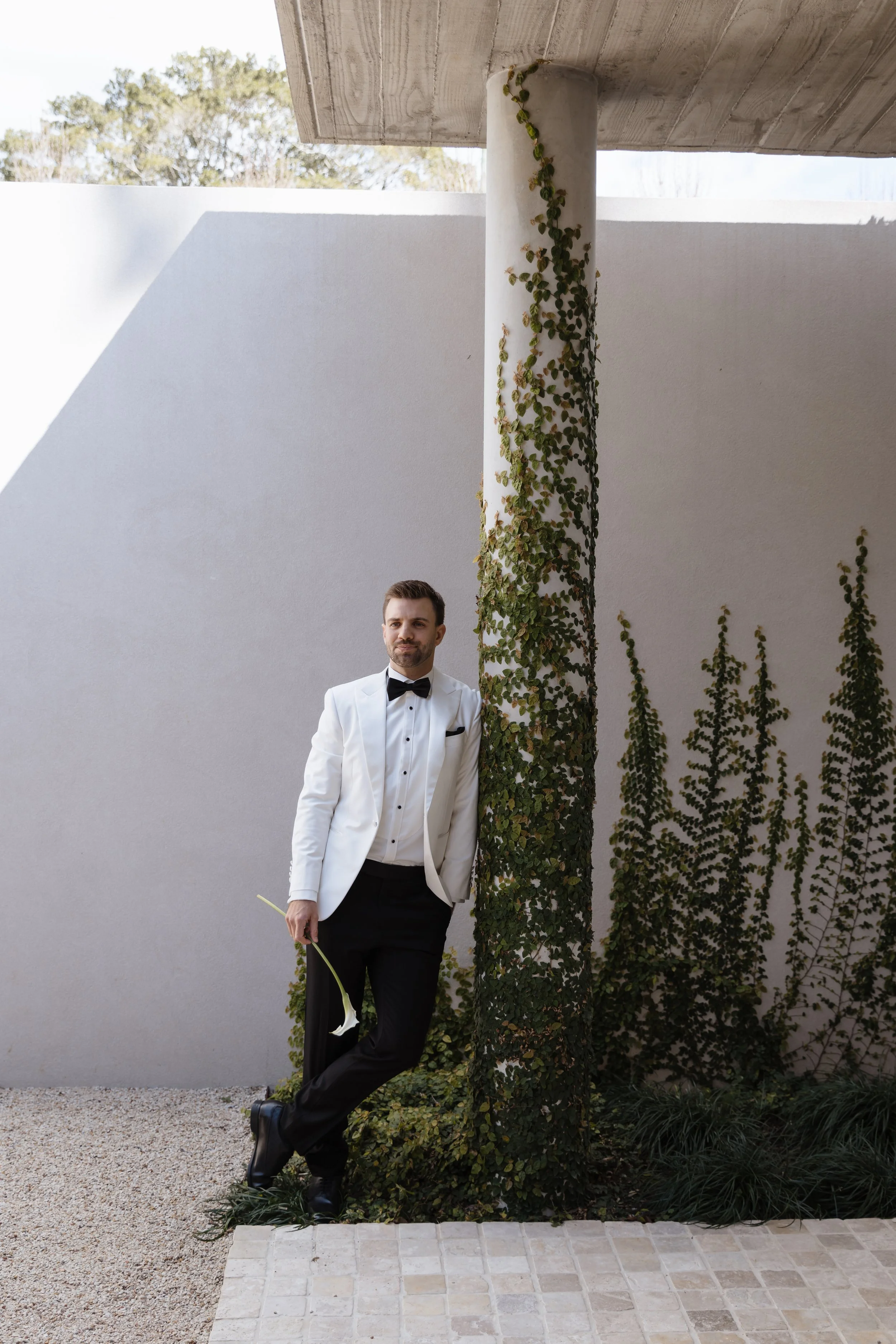 A man in a white tuxedo with a black bow tie, holding a white calla lily, standing against a background of a white wall, a concrete pillar covered in ivy, and some greenery, with a roof overhead.