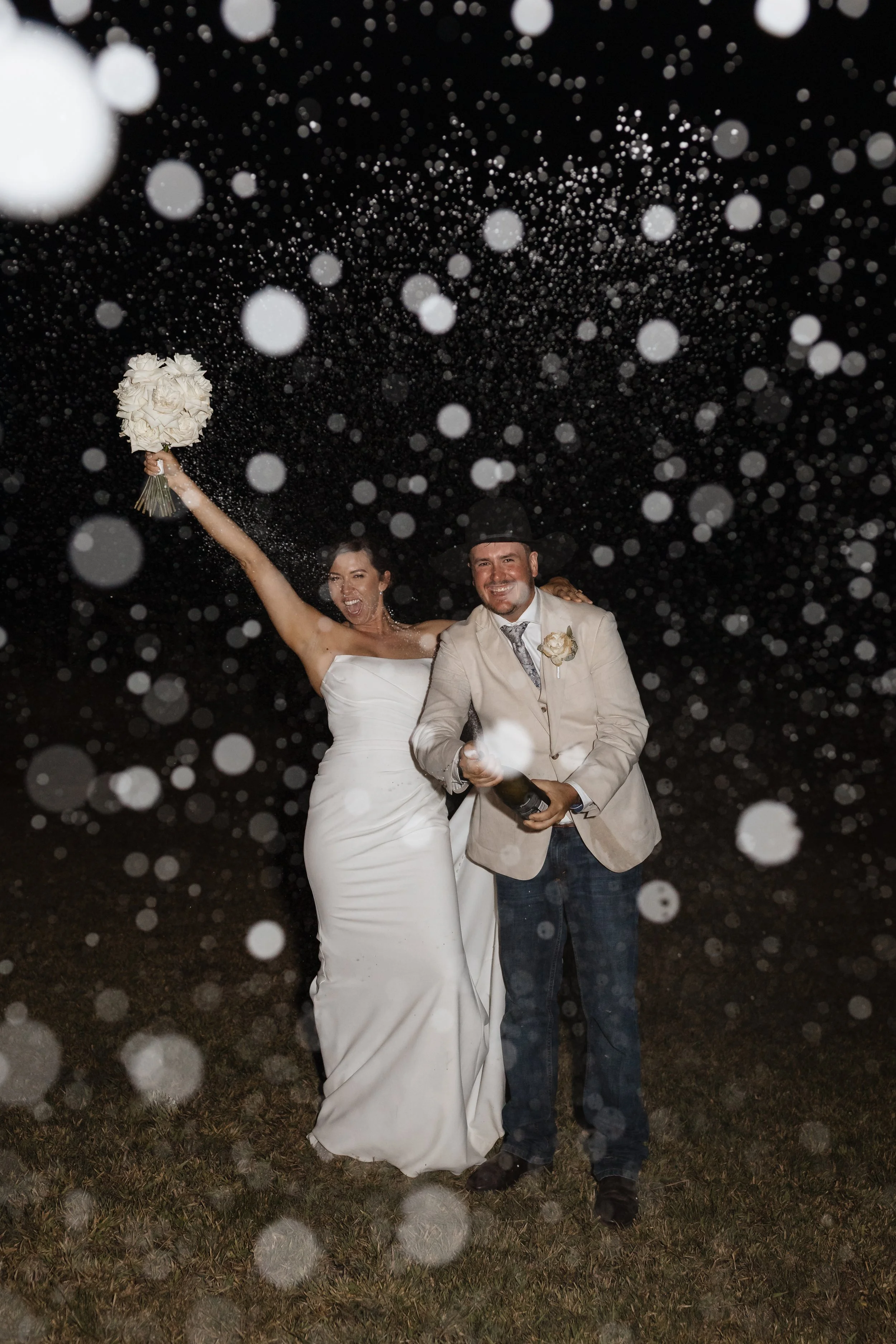 A newlywed couple celebrating outdoors at night, with rain or bubbles falling around them. The bride is wearing a white wedding dress and holding a bouquet, smiling and raising her arm. The groom is in a beige suit jacket, shirt, and tie, holding a b