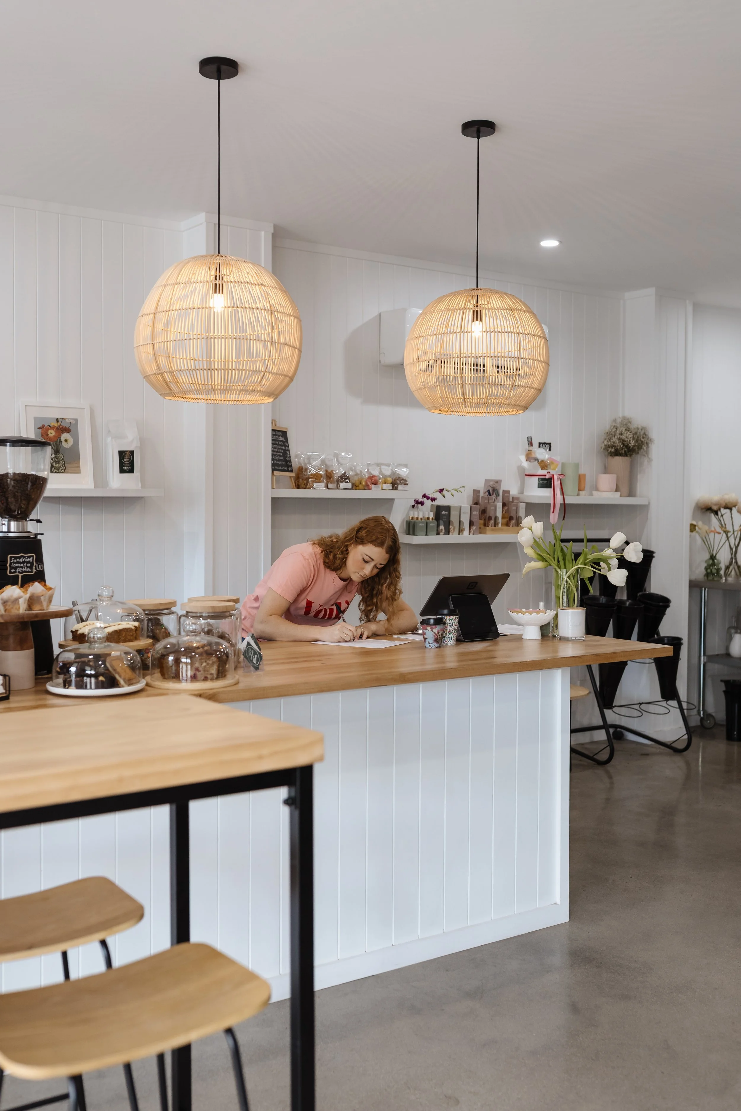 Inside a modern café with white paneled walls, two rattan pendant lights hanging from the ceiling, a woman in a pink shirt writing at the counter, and shelves with various products and decorations, including flowers and framed pictures.