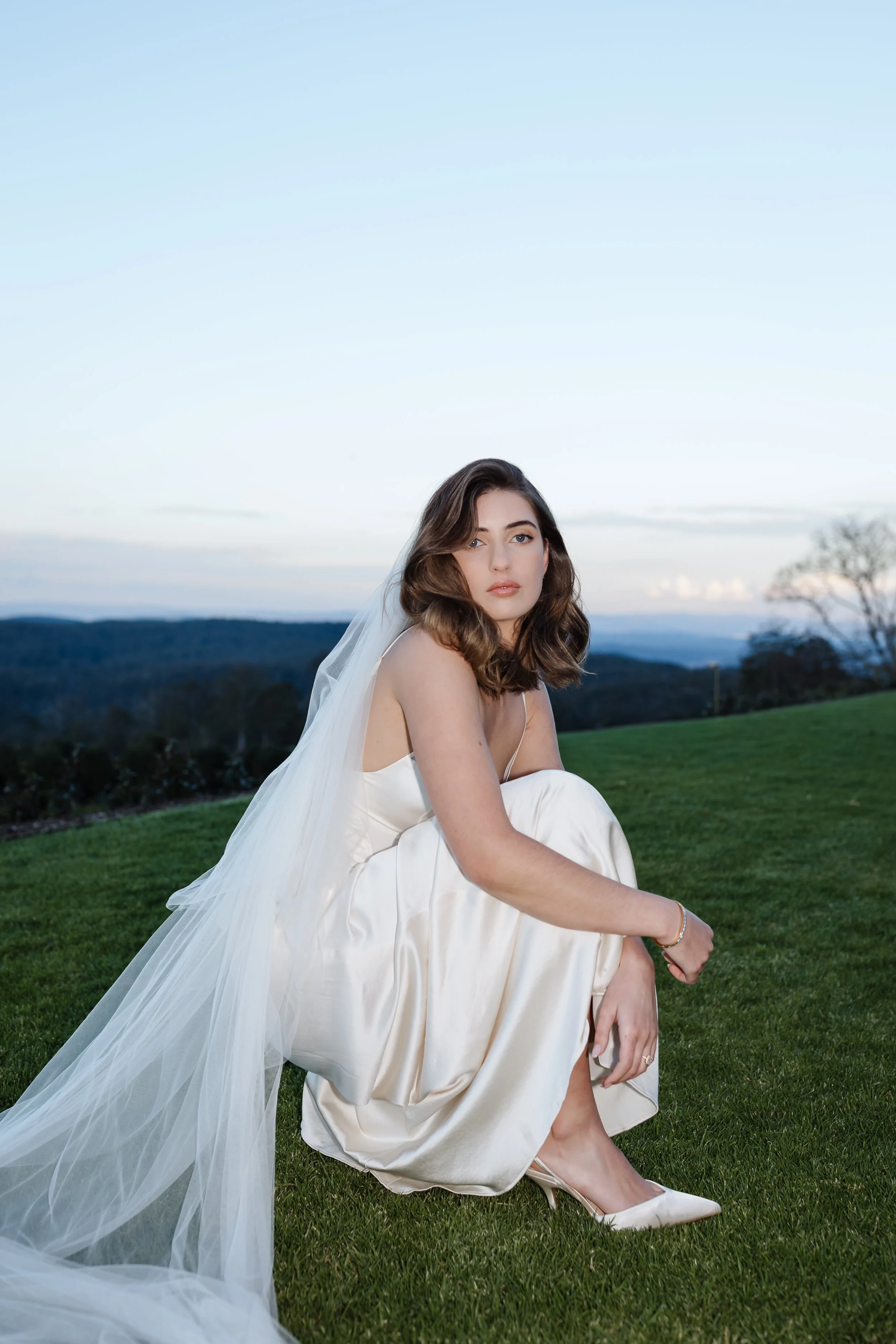 A woman in a white dress and veil sitting on a grassy field with mountains and a blue sky in the background.