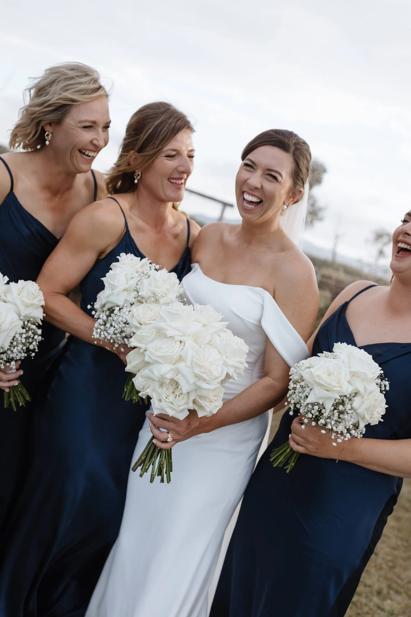 Five women dressed in wedding and bridesmaid dresses, smiling and laughing, holding bouquets of white roses and baby's breath, outdoor setting with cloudy sky and trees in the background.