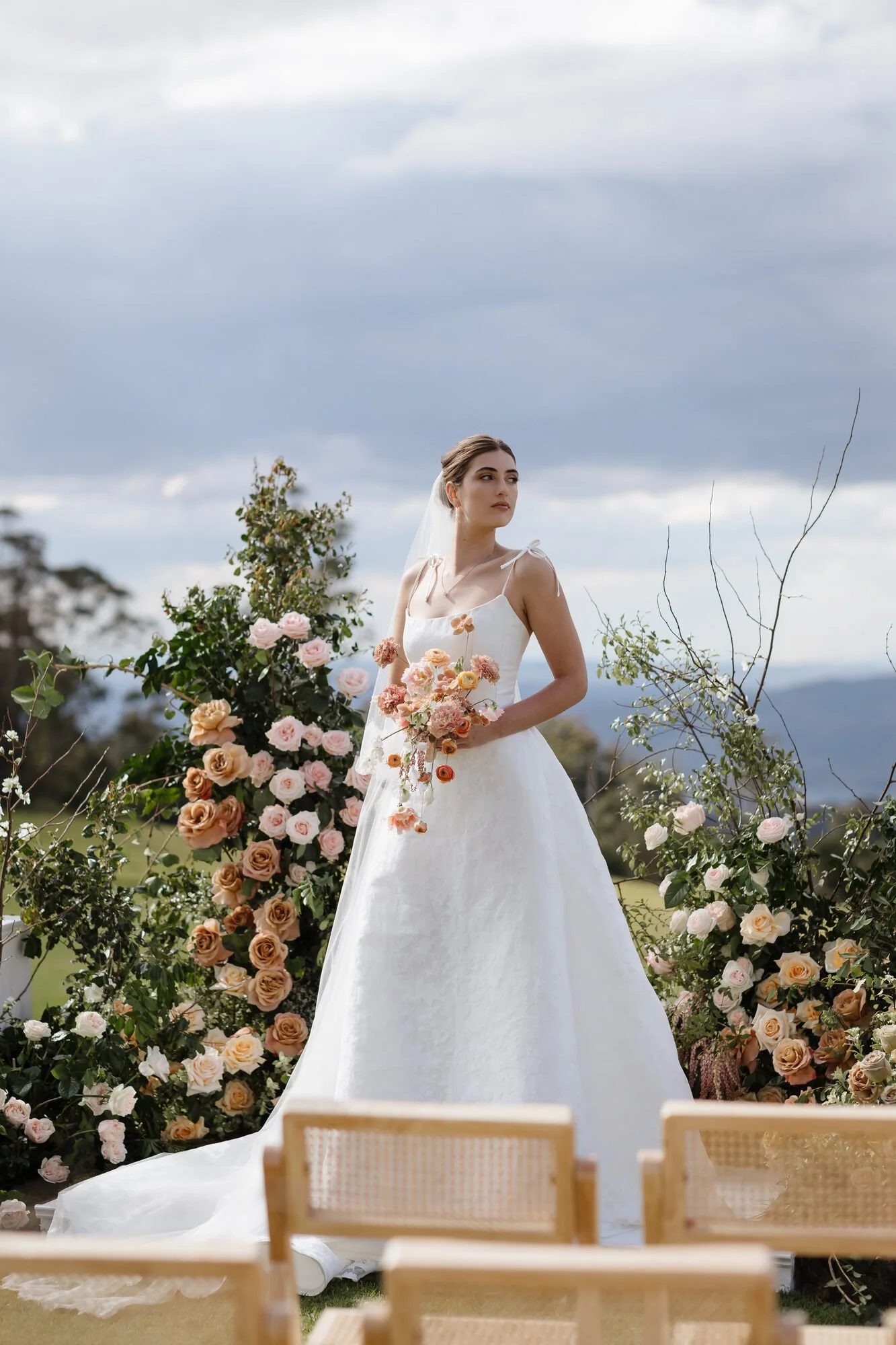A bride in a white wedding dress standing outdoors near a floral decoration with pink and peach roses, greenery, and branches, against a cloudy sky and distant mountains.
