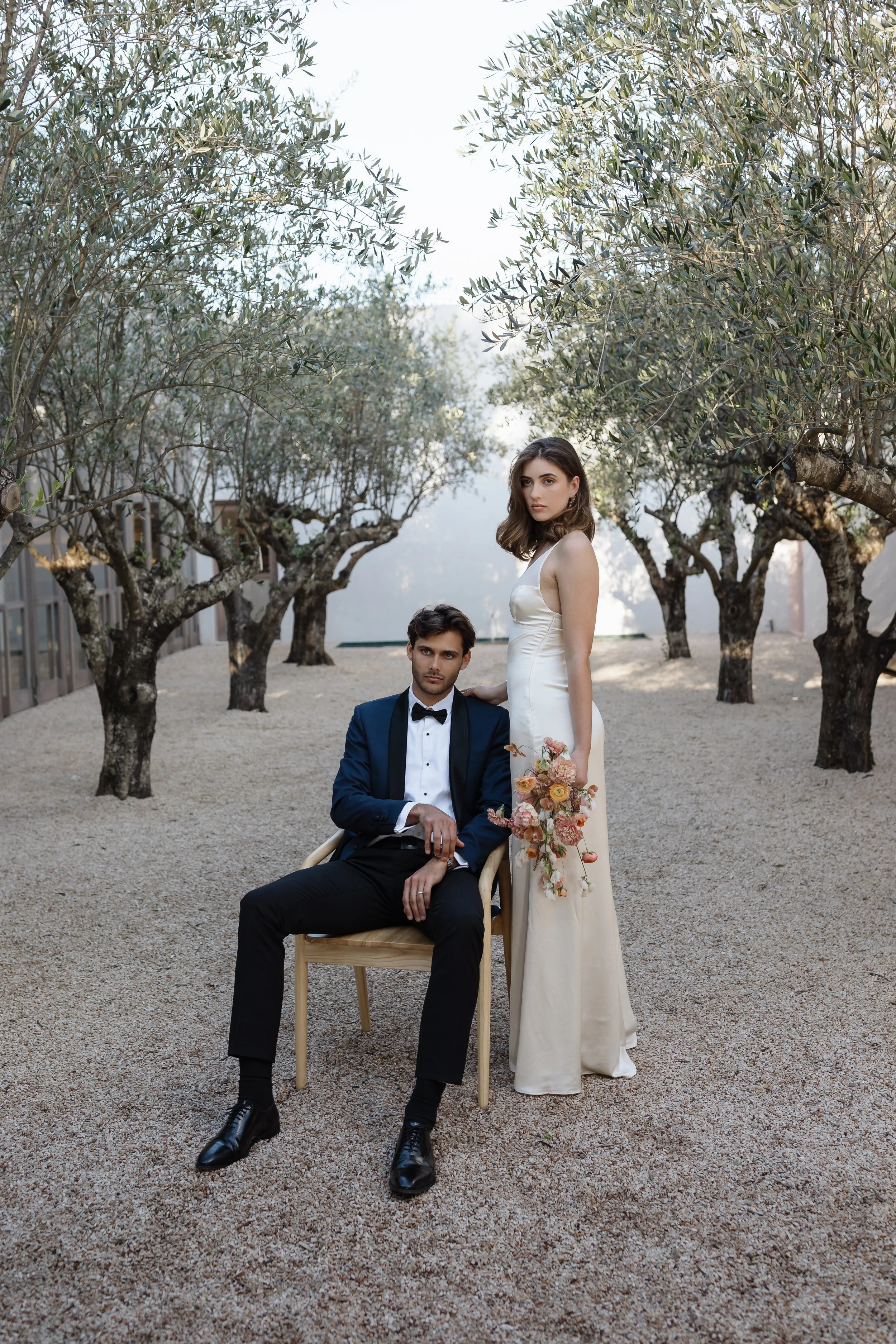 A man in a tuxedo sitting on a wooden chair with a woman in a white dress standing beside him, holding a bouquet of flowers, in an outdoor setting with trees and gravel ground.