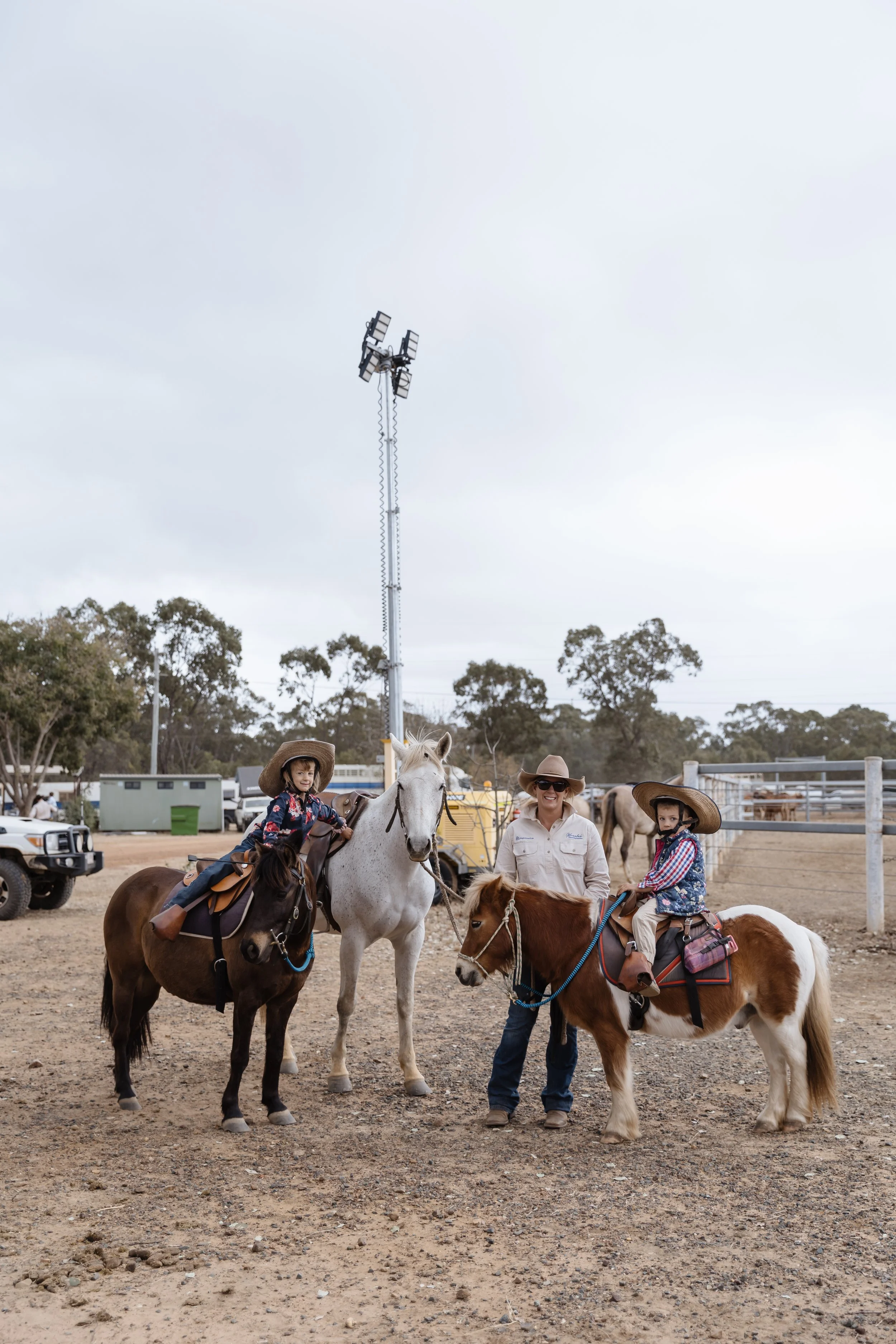 Two children riding horses with an adult woman standing between them at an outdoor equestrian event. All are wearing cowboy hats, and the children are dressed in western attire. There are other horses and people in the background, along with a tall l