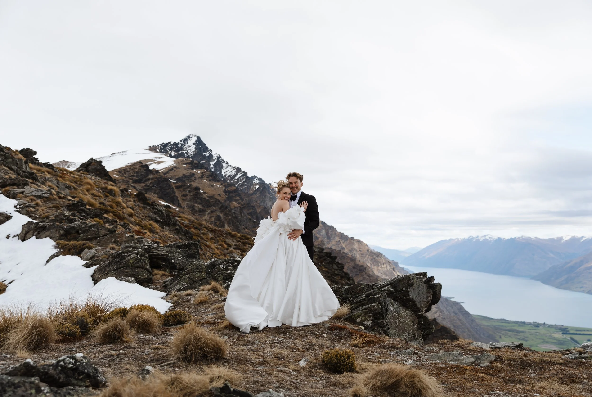 A couple in wedding attire standing on a rocky mountainside with snow patches, mountains, a lake, and an overcast sky in the background.