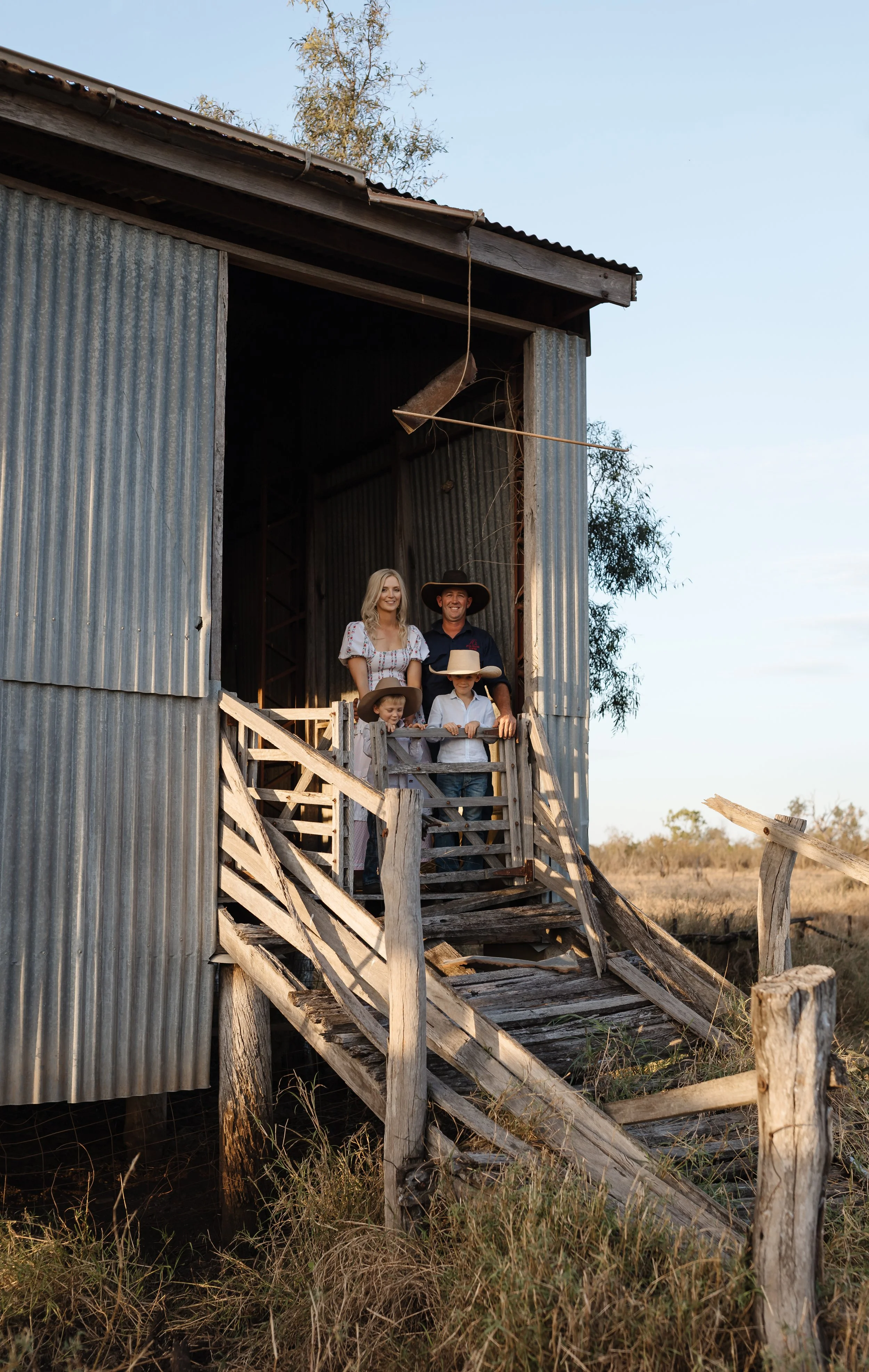 A family of five standing on the wooden stairs of an old barn with metal siding in a rural setting at sunset. The family includes a woman, a man, and three children, all dressed in casual western-style clothing, with two children wearing cowboy hats.