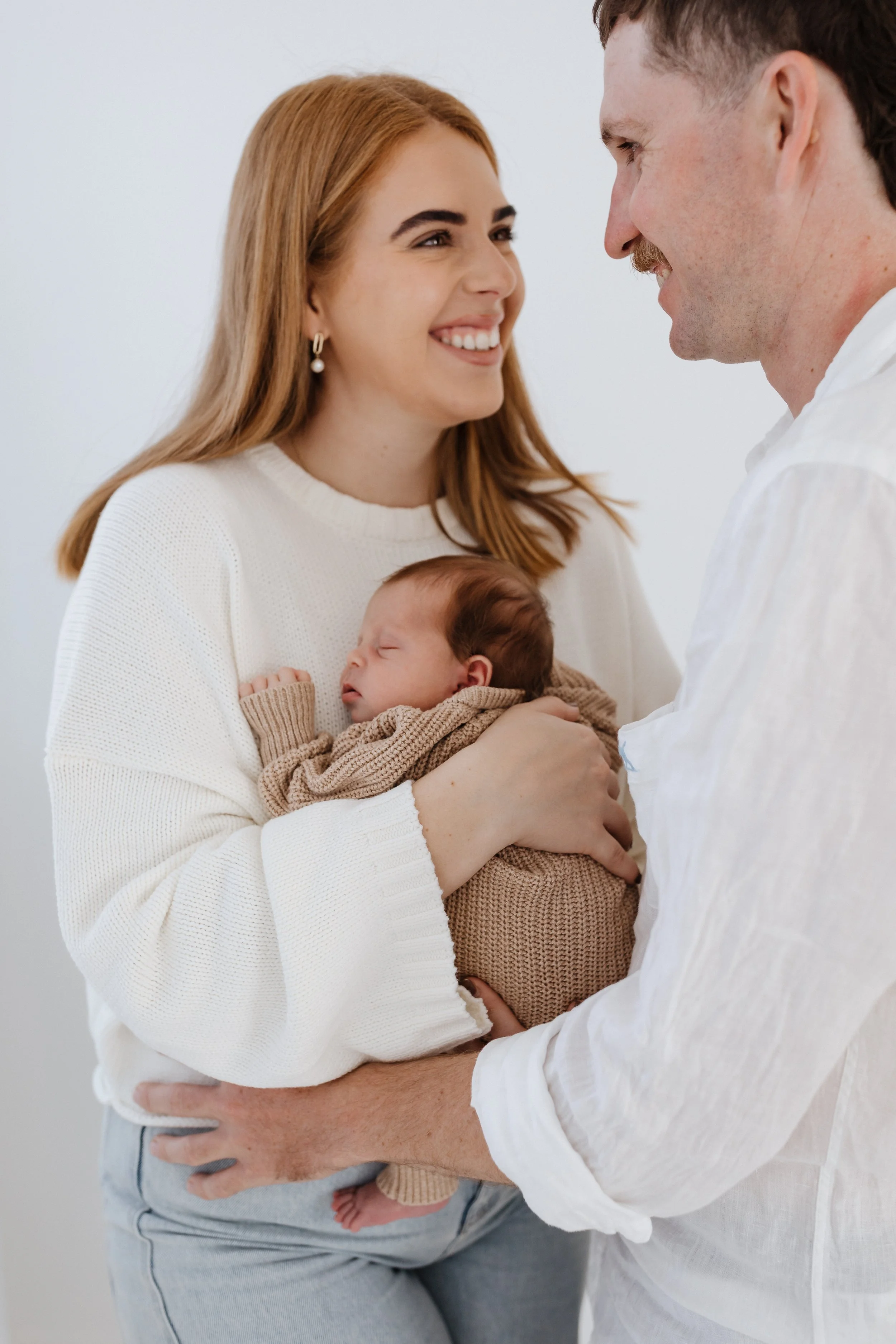 A woman with red hair holding a sleeping baby, while a man with dark hair and a smile faces them and touches the woman's arm. The scene is warm and loving, likely in a home or health setting.
